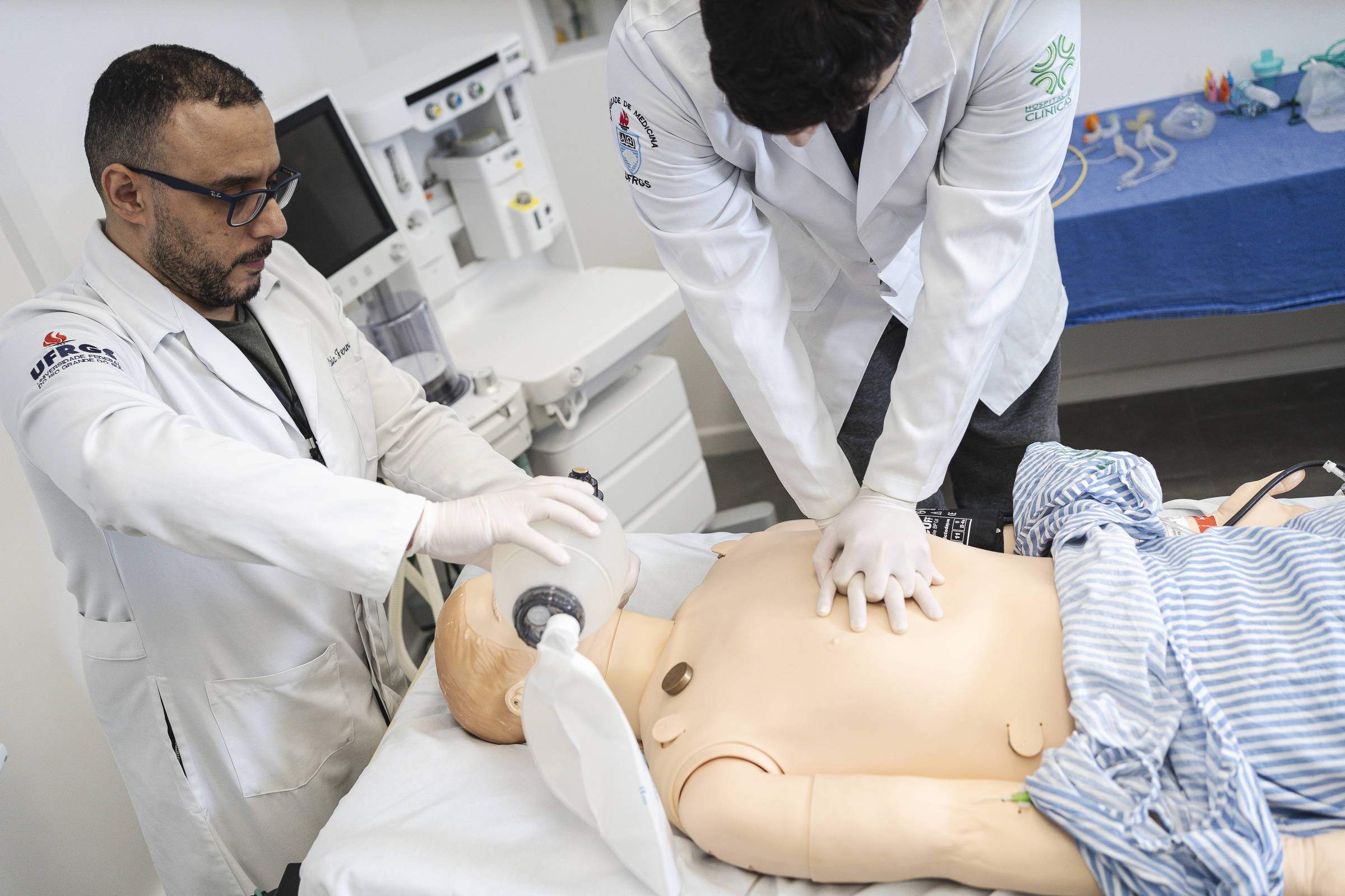Dois profissionais de saúde em jalecos brancos realizam treinamento de reanimação cardiopulmonar em manequim adulto sobre maca. Um aplica compressões torácicas enquanto o outro utiliza bolsa de ventilação manual.