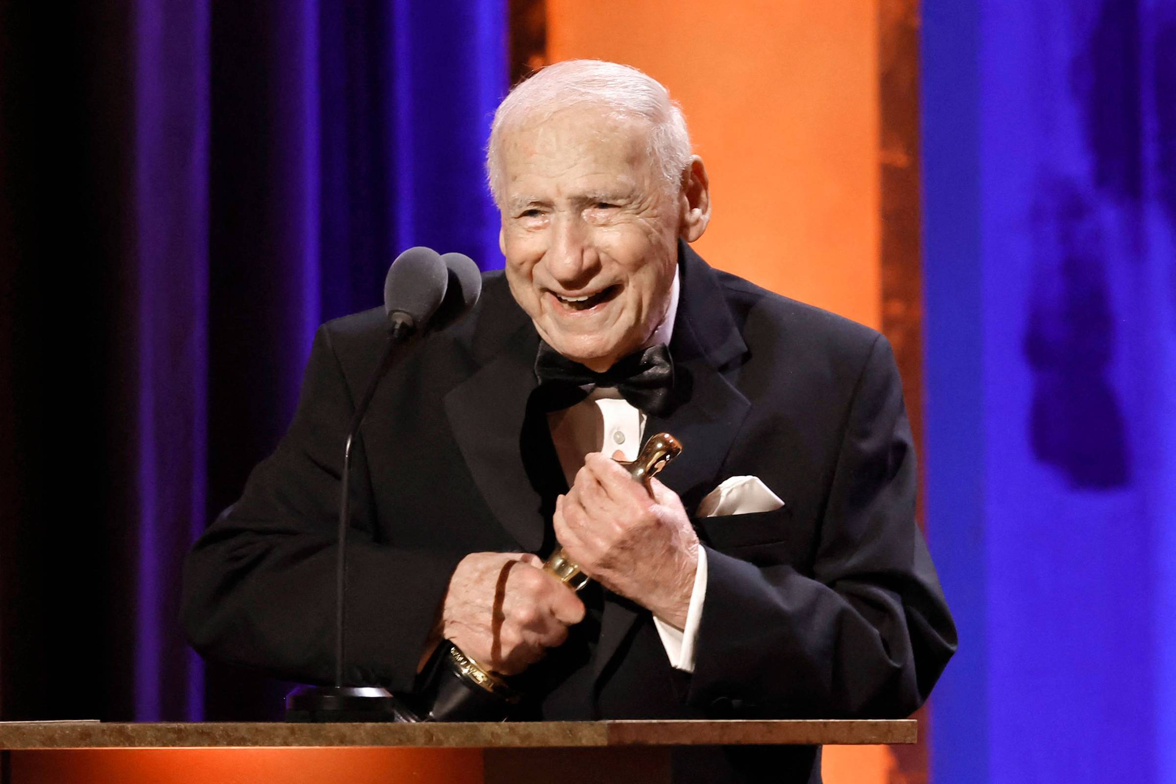 Homem idoso de cabelo branco veste smoking preto com gravata borboleta e segura estatueta dourada em cerimônia. Ele está atrás de um púlpito com microfone, com fundo iluminado em tons de azul e laranja.