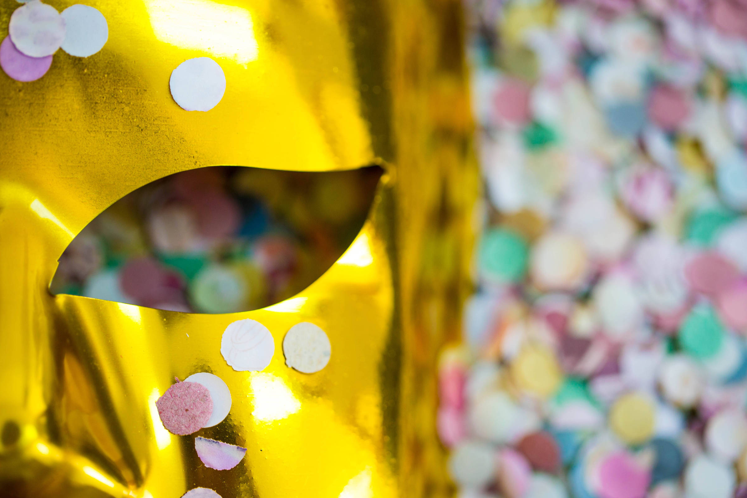 A imagem mostra uma máscara dourada com um recorte em forma de olho. A máscara está decorada com pequenos confetes coloridos, incluindo círculos em rosa, branco e azul. Ao fundo, há uma superfície coberta por confetes em várias cores, criando um ambiente festivo.
