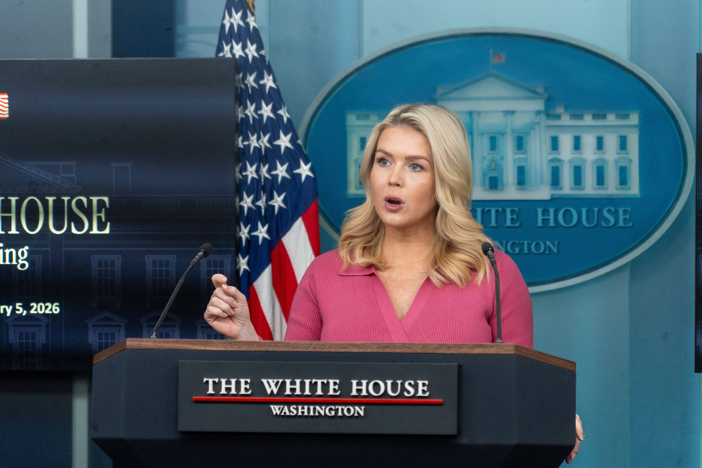 Mulher loira fala em púlpito da Casa Branca com bandeira dos EUA ao fundo e logo oficial visível.