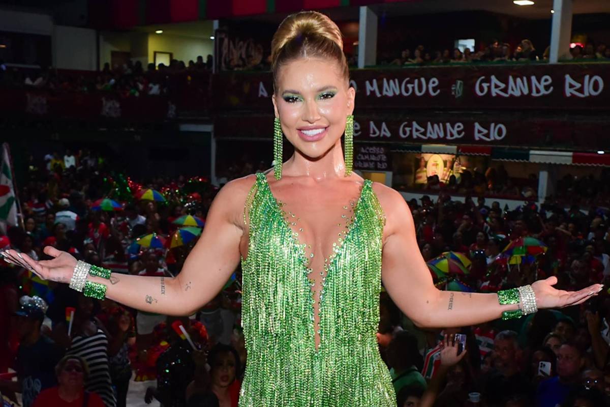 Mulher sorridente com cabelo preso em coque alto usa fantasia verde com franjas brilhantes e acessórios combinando, posando com braços abertos diante de multidão em ambiente interno decorado para carnaval.