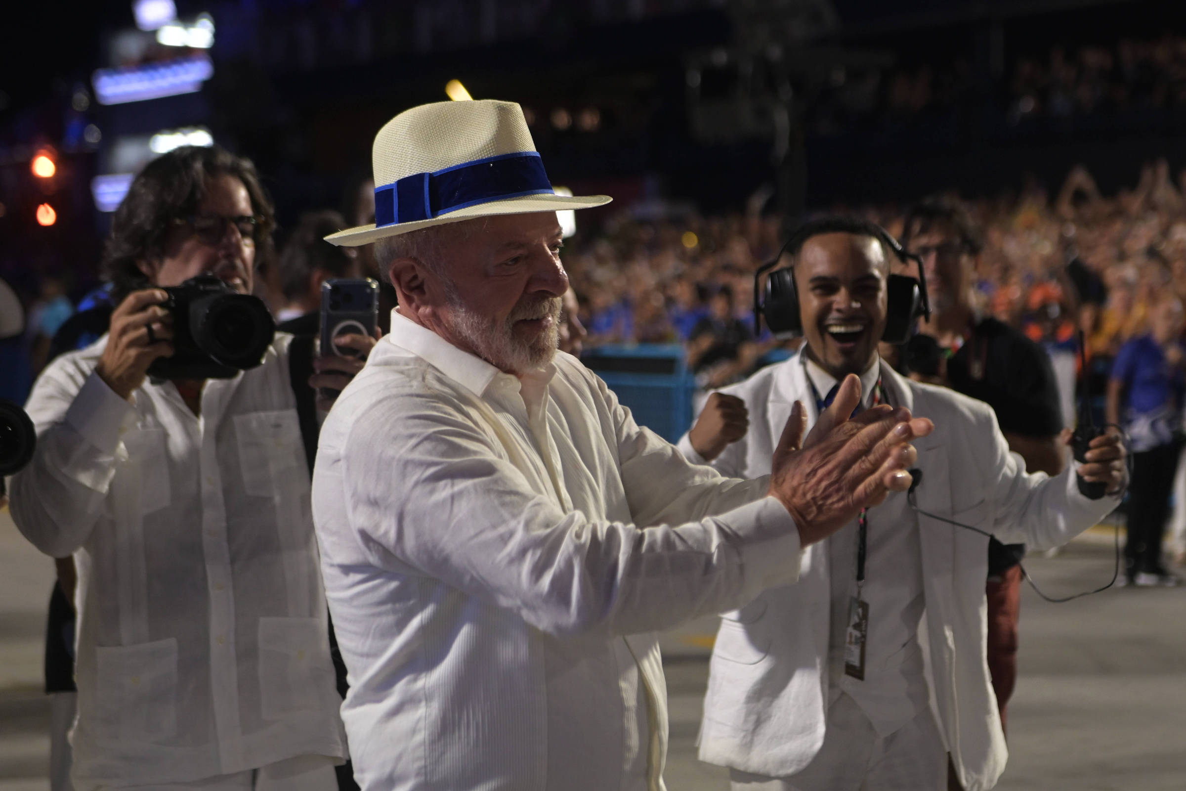 Homem idoso de chapéu branco com faixa azul e camisa branca aplaude em primeiro plano. Atrás dele, homem com câmera fotográfica e outro com fones de ouvido sorriem. Multidão desfocada ao fundo em ambiente iluminado.