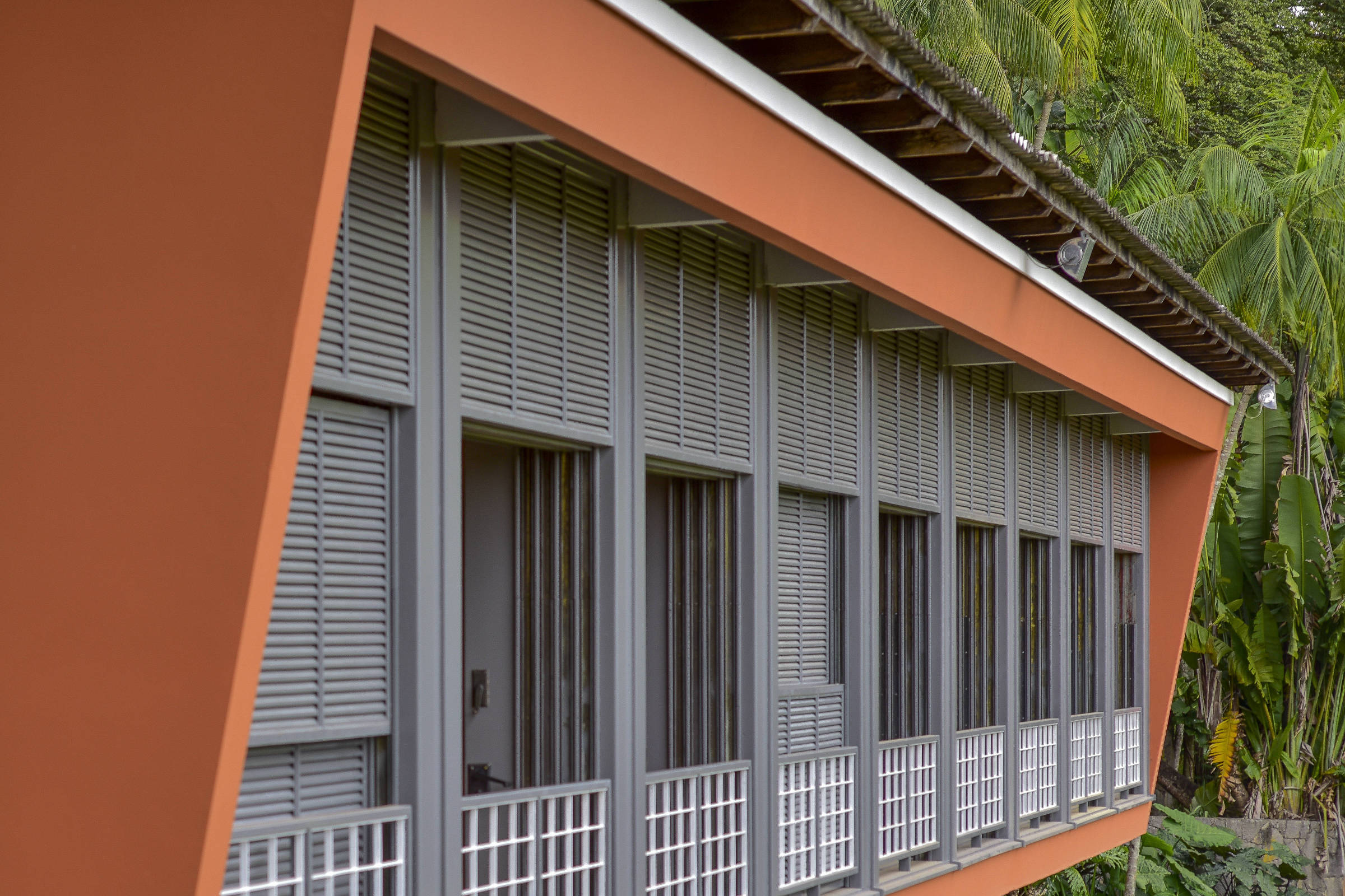 Prédio com fachada lateral laranja e janelas cinza com persianas fechadas, sustentado por pilares. Parede amarela com luminária à esquerda. Vegetação e pessoa sentada ao fundo.