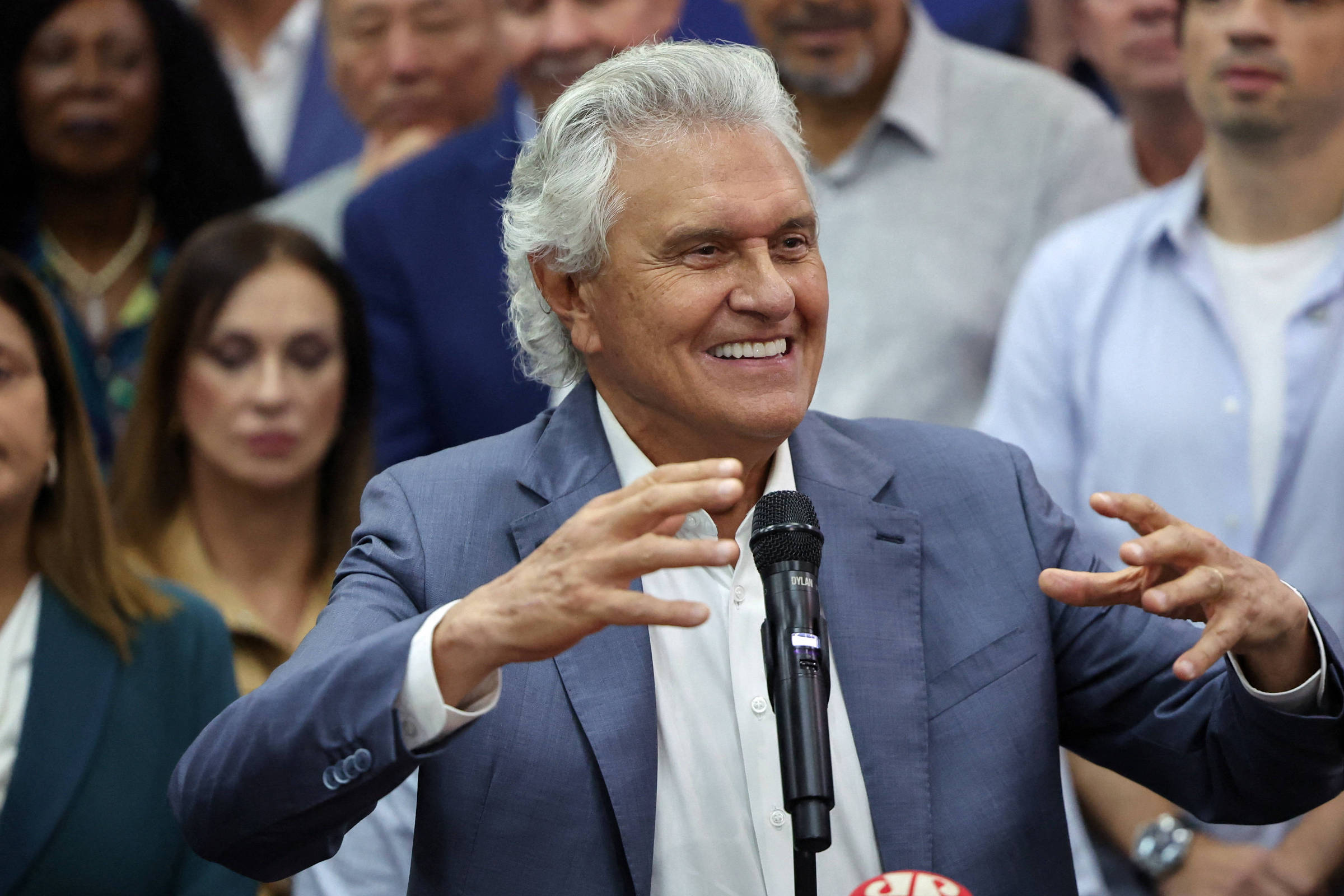Homem de cabelos grisalhos e terno azul fala gesticulando em microfone durante coletiva. Pessoas diversas observam ao fundo.