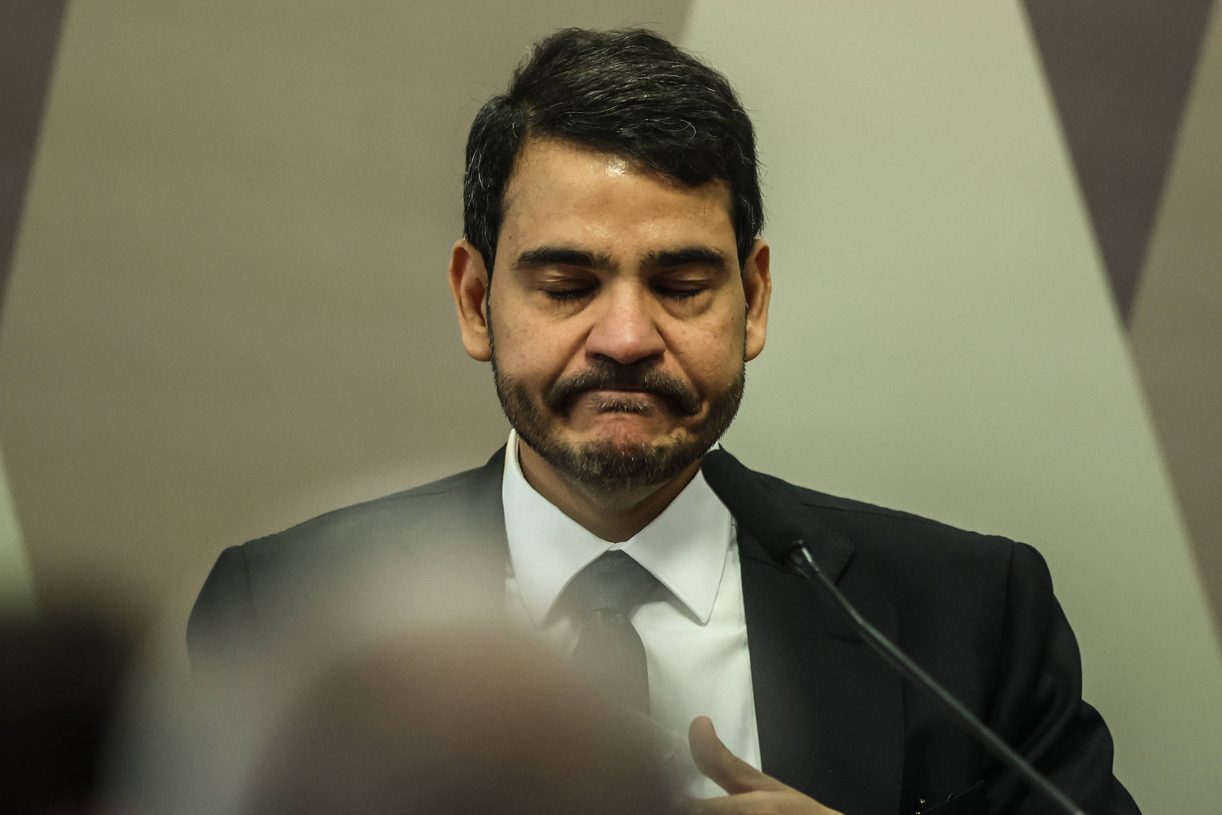 Homem com cabelo curto e barba aparada veste terno preto, camisa branca e gravata preta, com expressão séria e olhos fechados, em frente a microfone.