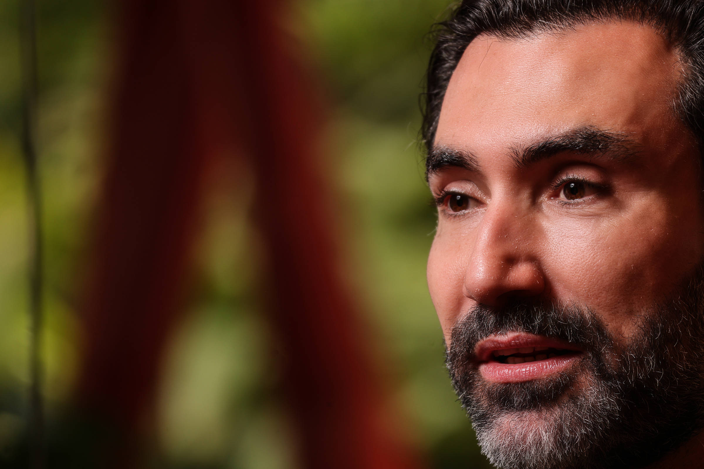 Homem de pele clara com barba e cabelo escuro, olhando levemente para a direita. Fundo desfocado em tons verdes e vermelhos.