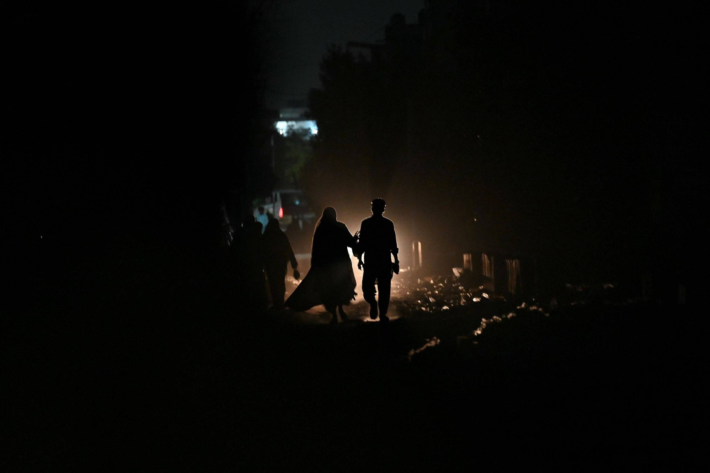 Duas pessoas caminham de mãos dadas em uma área escura, iluminadas por uma luz forte atrás delas que cria silhuetas nítidas. O entorno é pouco visível, com sombras e objetos indistintos ao redor.