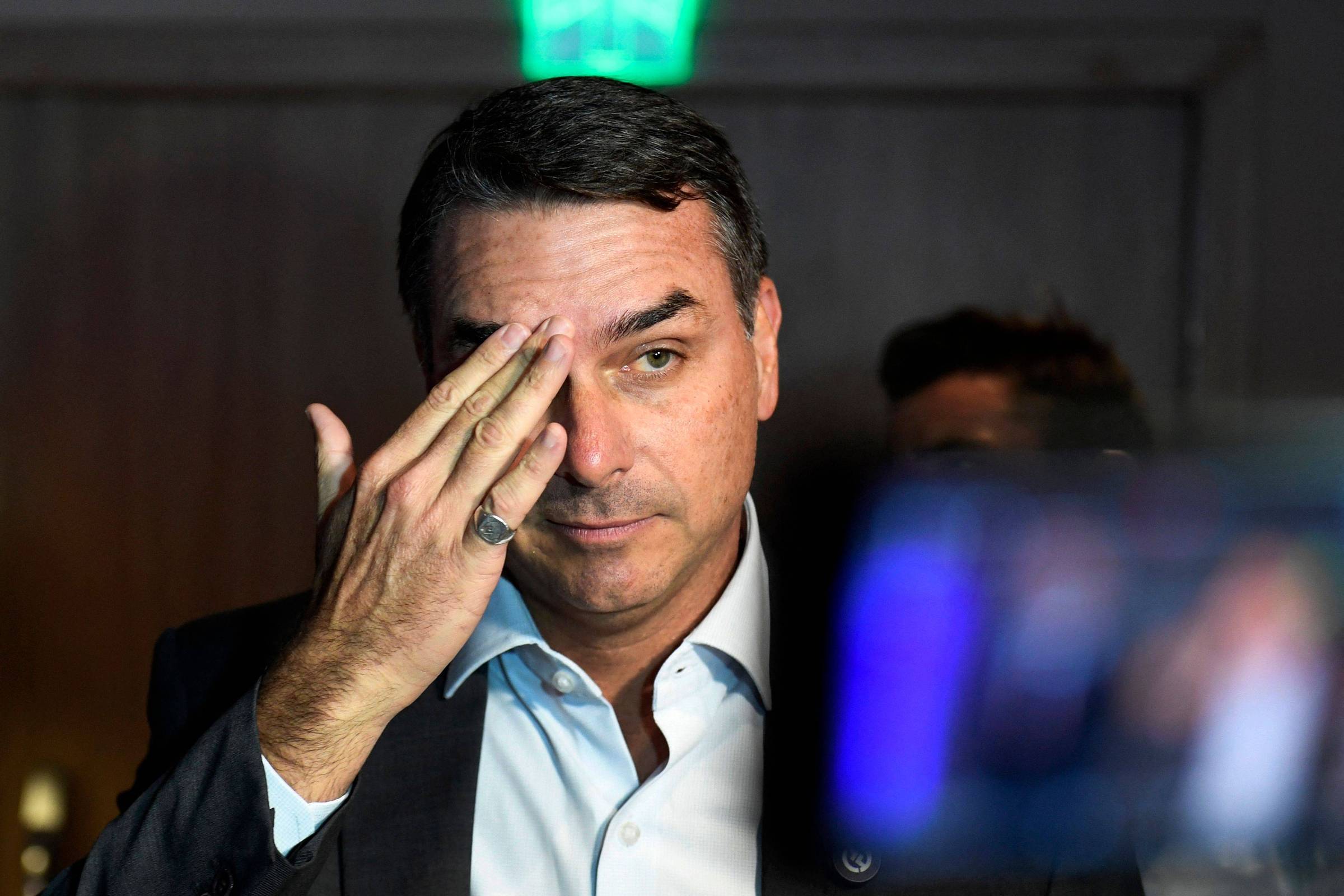 Homem de meia-idade com cabelo curto e escuro, veste camisa branca e paletó escuro, ajusta o rosto com a mão esquerda, em ambiente interno com fundo desfocado e luz verde ao fundo.