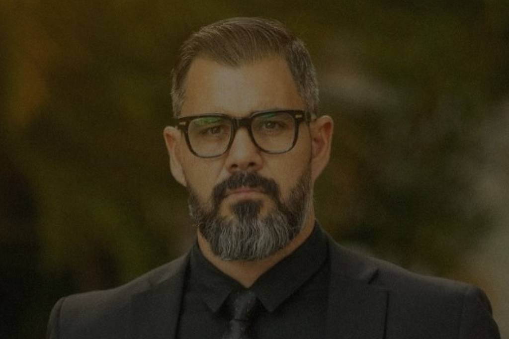 Homem de barba e óculos escuros veste terno preto e gravata cinza, posando ao ar livre com fundo desfocado. Texto sobreposto diz: