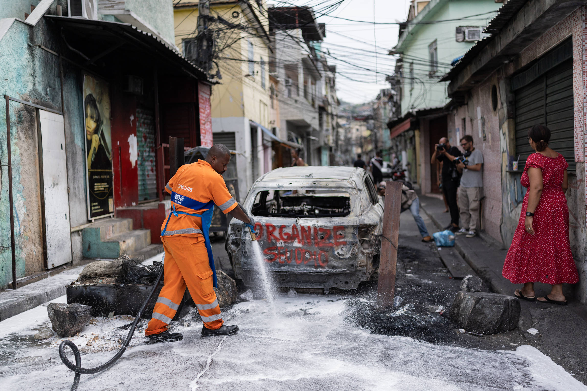 Homem com uniforme laranja usa mangueira para lavar rua queimada em área residencial. Carro destruído pelo fogo com grafite