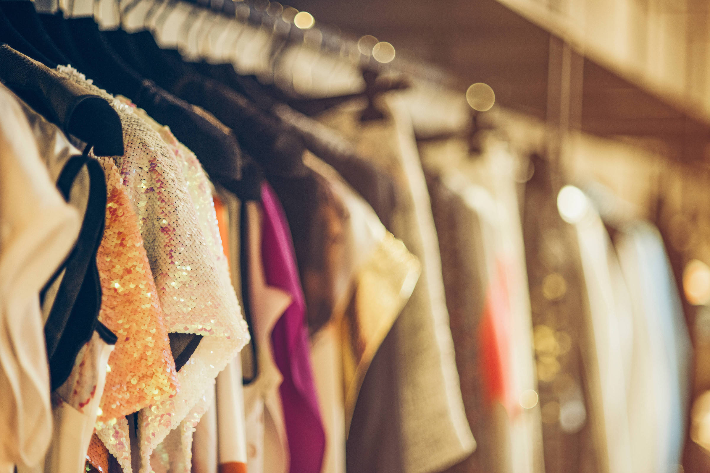 A imagem mostra uma variedade de roupas penduradas em cabides. As peças incluem blusas e vestidos, com diferentes texturas e cores, como um vestido brilhante em tons de laranja e outras roupas em tons neutros e vibrantes. O fundo é desfocado, sugerindo um ambiente de loja ou boutique.