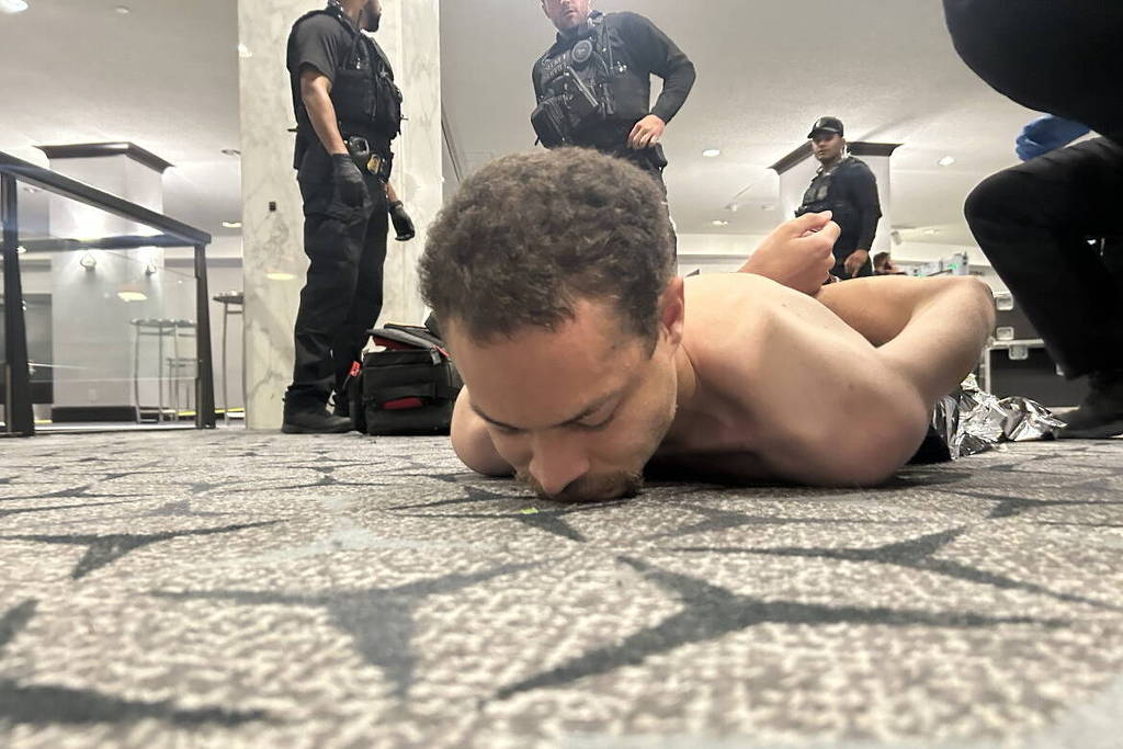 Homem sem camisa está deitado de bruços no chão com as mãos algemadas atrás das costas. Três policiais em uniforme preto estão em pé ao fundo em ambiente interno com piso de carpete.