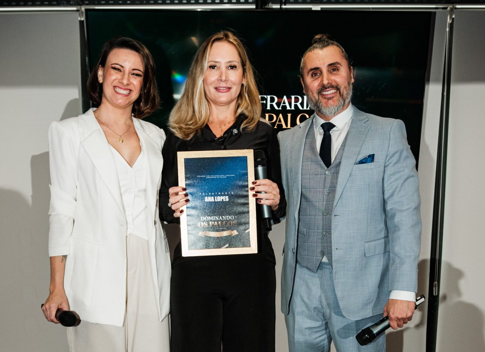 Ana Lopes segura placa de reconhecimento durante evento da Confraria dos Palcos ao lado de Bruna Grah e Fernando Freitas em palco de premiação.
