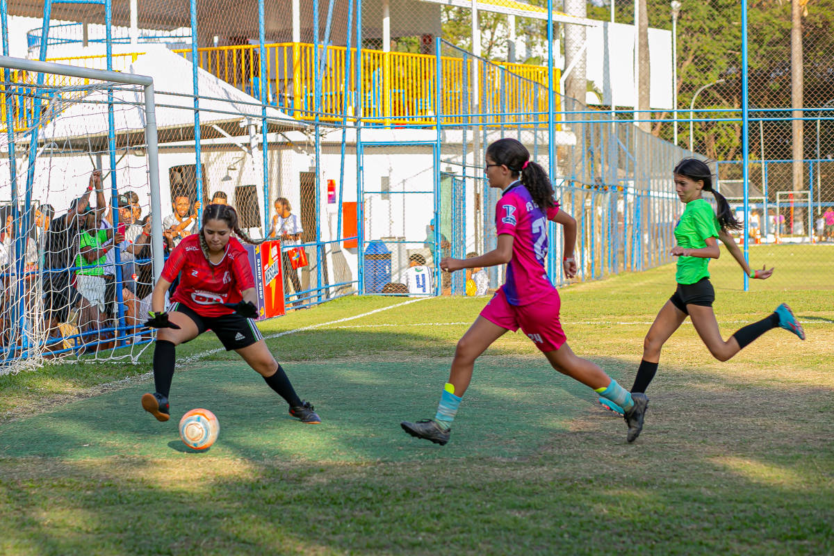 Meninas estão em um campo de futebol jogando; detalhe de um chute a gol em que a goleira pega a bola; ao fundo se vê vários torcedores