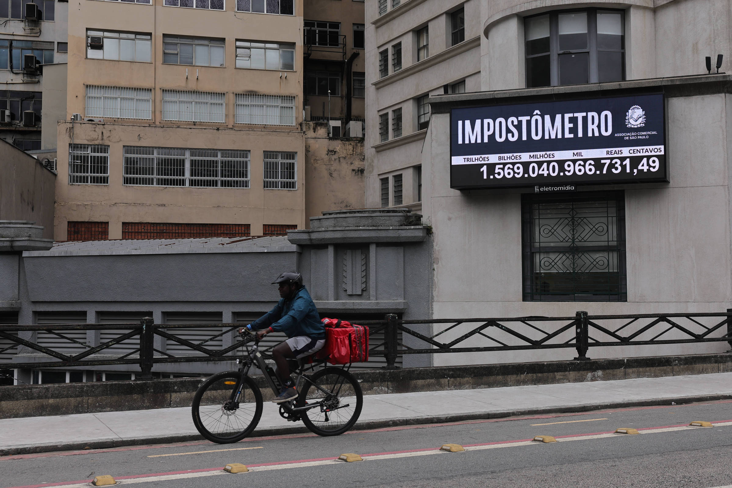 Impostômetro digital exibe valor acumulado de impostos em fachada de prédio antigo. Ciclista com mochila vermelha pedala na ciclovia em primeiro plano, com edifícios residenciais ao fundo.