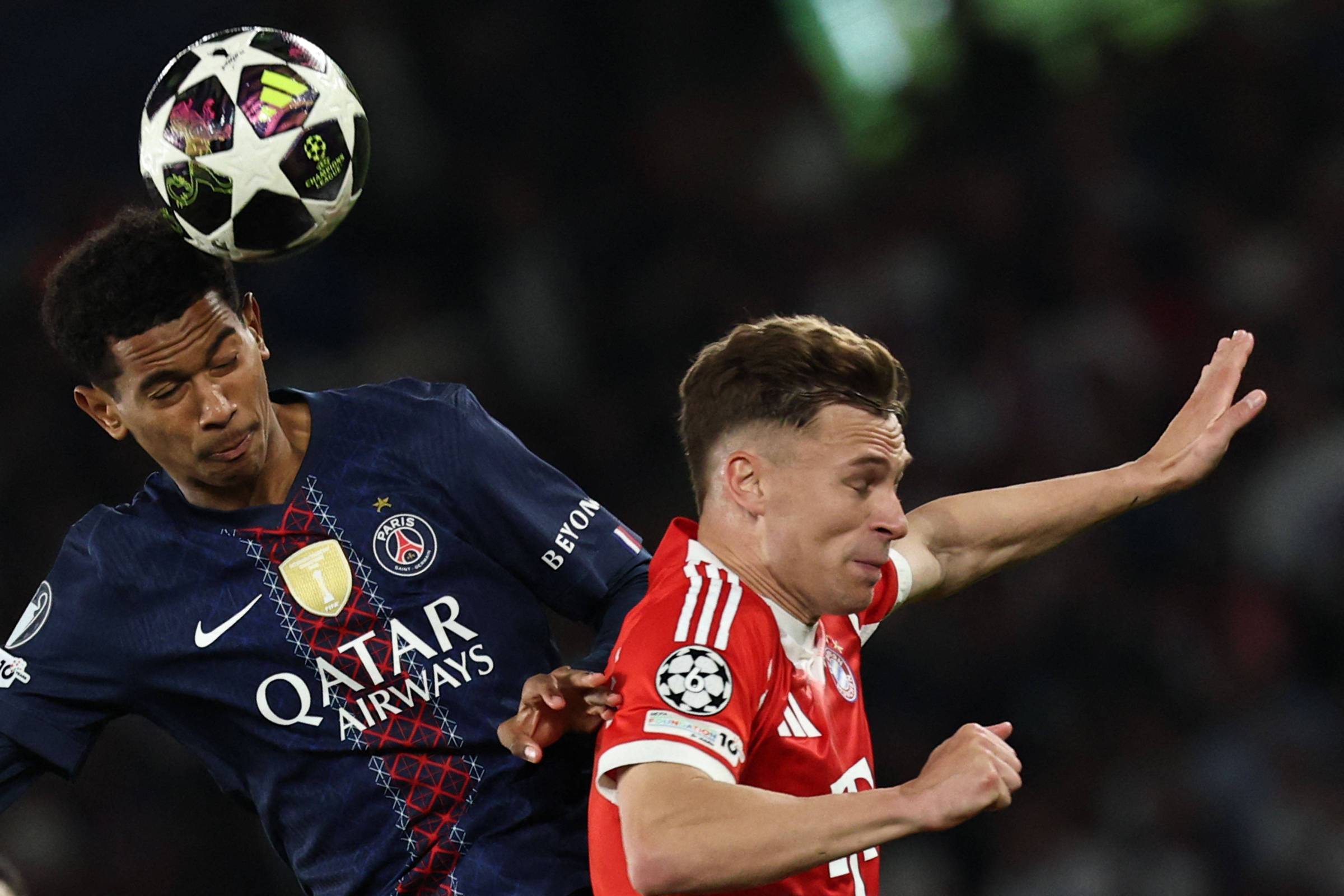 Dois jogadores disputam a bola pelo alto durante partida noturna da Liga dos Campeões. Jogador do PSG veste uniforme azul escuro e jogador do Bayern de Munique veste uniforme vermelho.