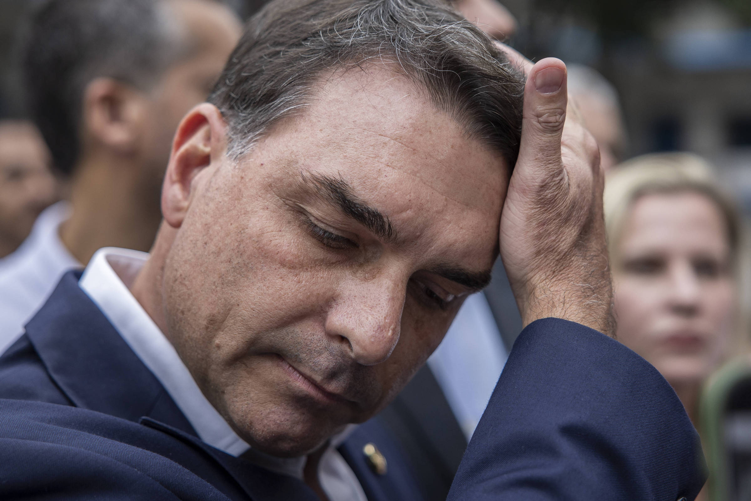Homem de terno azul escuro ajeita o cabelo com a mão direita, com expressão séria. Pessoas desfocadas aparecem ao fundo em ambiente externo.