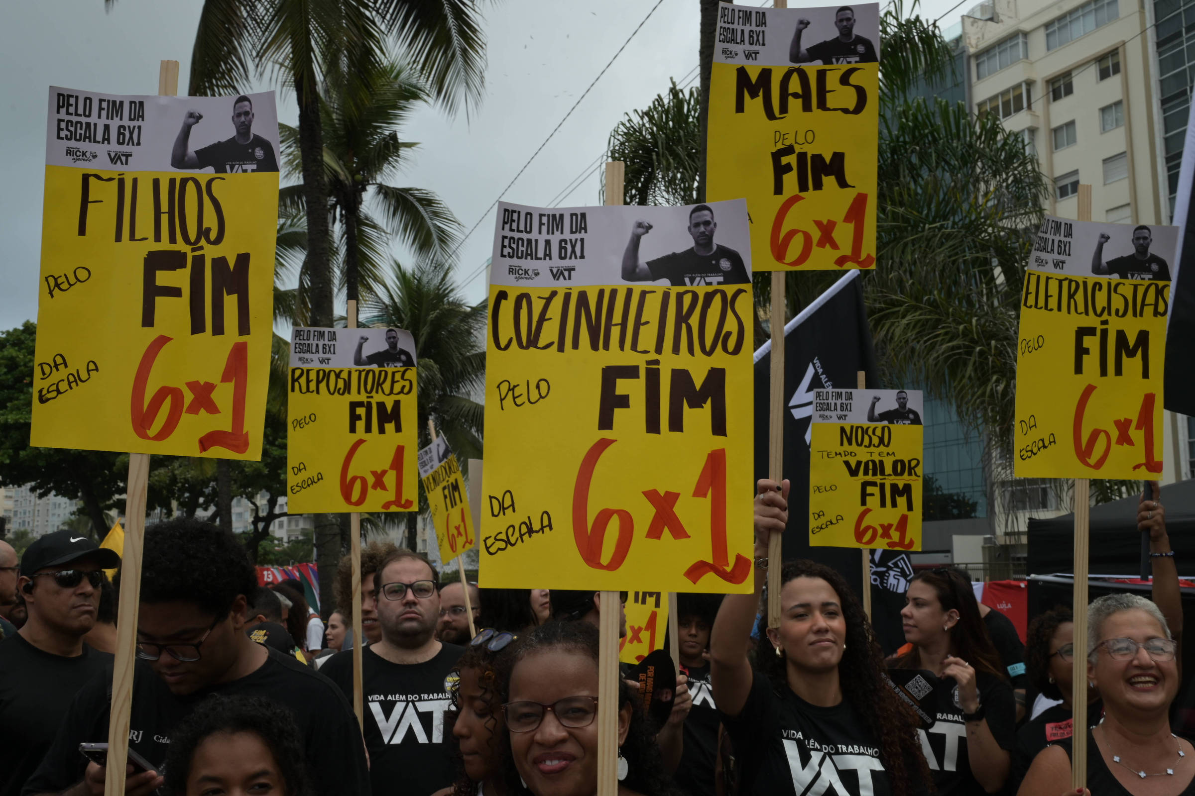 Grupo de manifestantes em rua urbana segura cartazes amarelos pedindo fim da escala 6x1 para diversas categorias, como filhos, cozinheiros, eletricistas e repositores. Pessoas vestem camisetas pretas com texto branco e há palmeiras e prédios ao fundo.