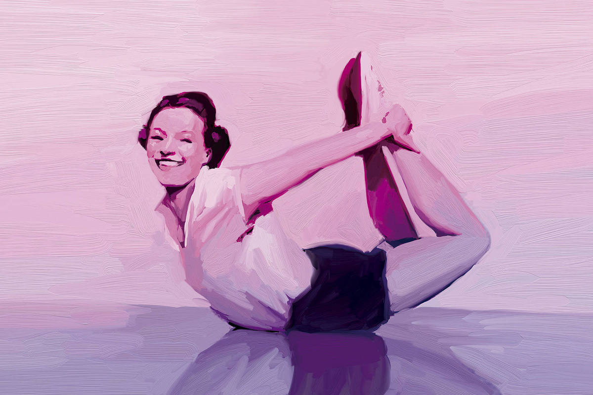 Mulher sorridente de cabelos curtos presos, vestindo camiseta branca e shorts escuros, realiza exercício de alongamento no chão, segurando os pés com as mãos atrás do corpo, com fundo em tons de rosa.