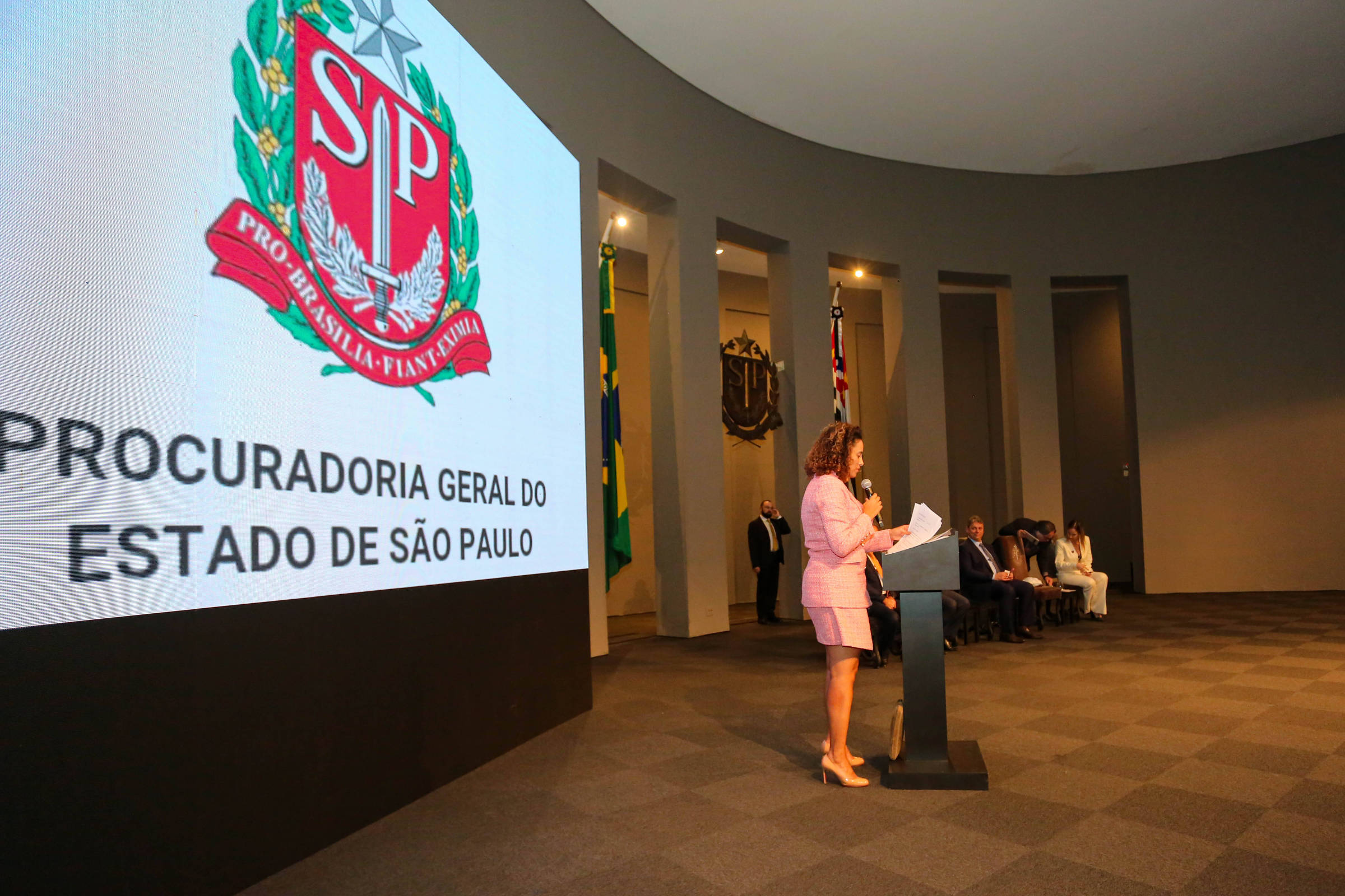 Uma mulher negra de pele clara falando ao púlpito em uma grande sala com um telão onde se lê Procuradoria Geral do Estado de São Paulo.