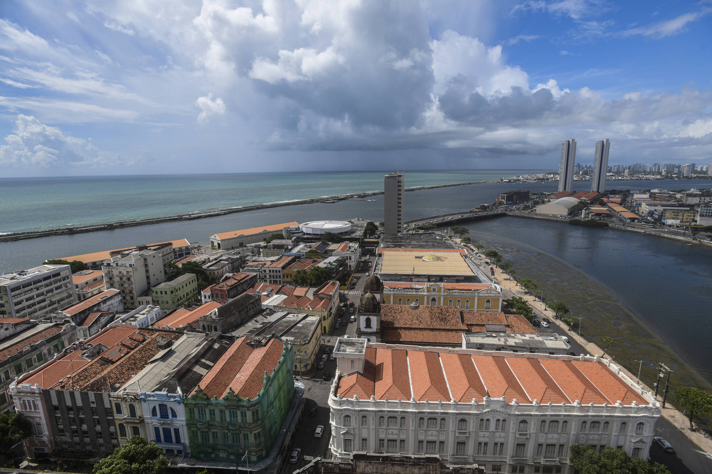 Vista aérea do centro histórico de Recife com prédios antigos de telhados vermelhos em primeiro plano, rio à direita e mar ao fundo sob céu parcialmente nublado.