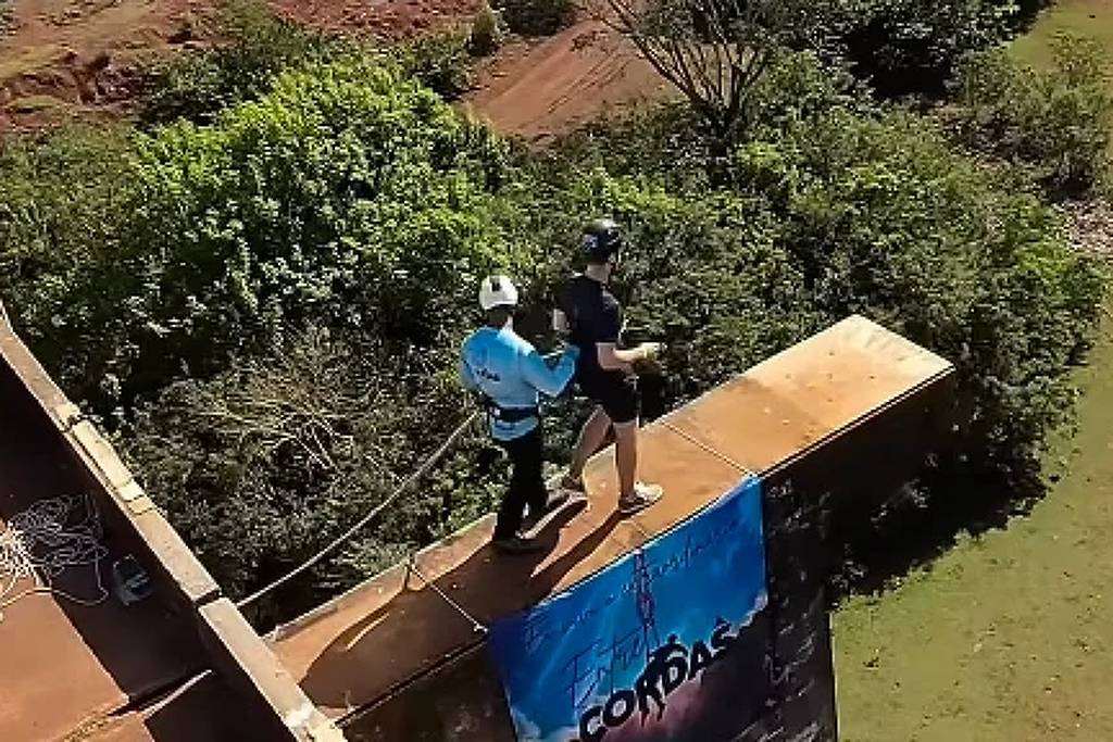 Duas pessoas estão em uma prancha de salto de madeira suspensa sobre uma estrutura metálica azul, cercada por vegetação e área de terra ao redor.