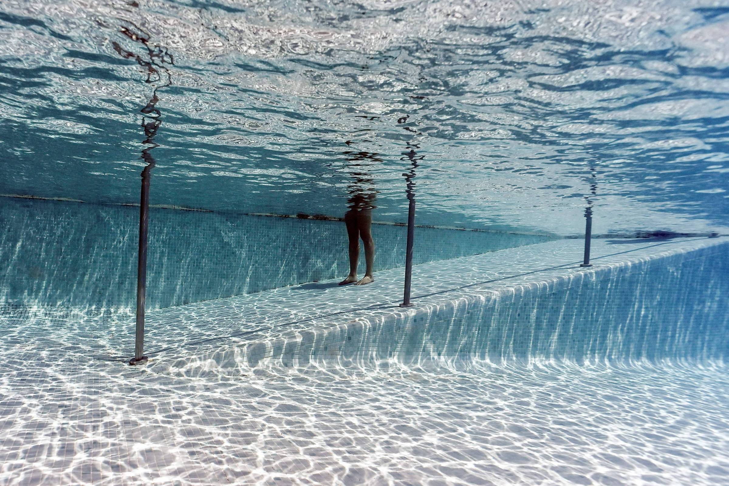 Duas pessoas caminham sobre passarela submersa em piscina com água clara. Estrutura metálica suporta passarela, e reflexos de luz criam padrões no fundo da piscina.