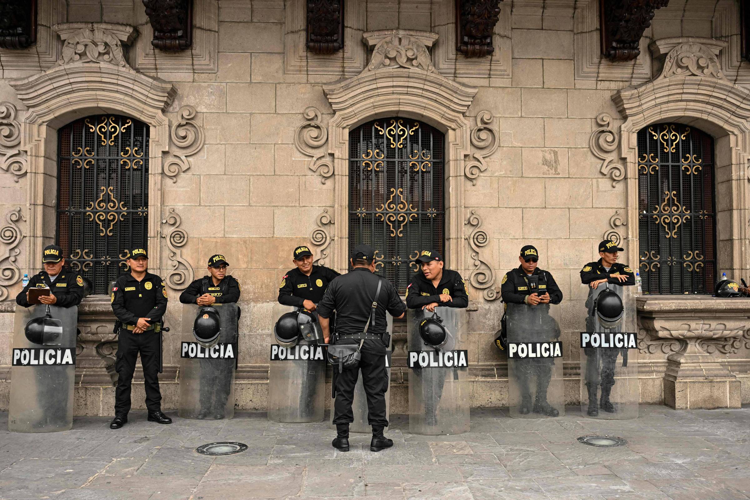 Oito policiais em uniforme preto formam linha em frente a um prédio de pedra com janelas ornamentadas. Sete seguram escudos transparentes com a palavra