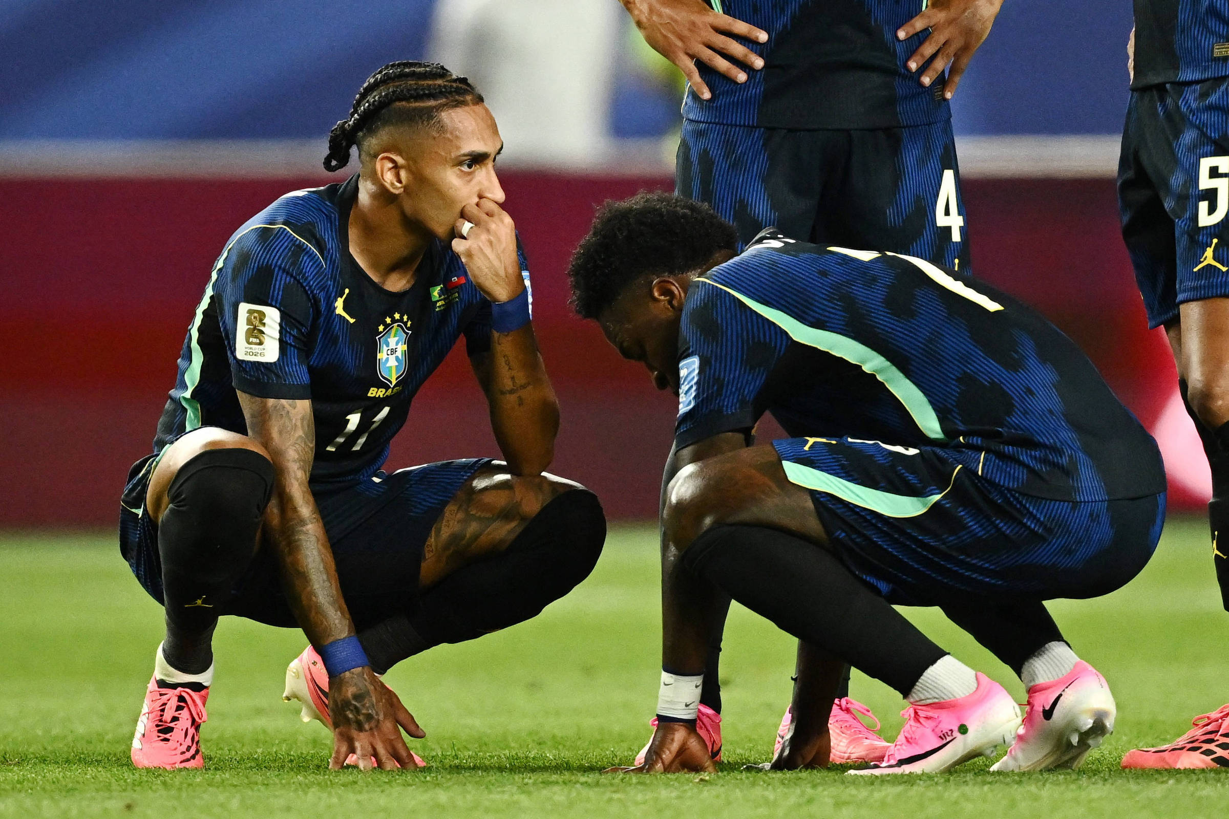 Dois jogadores da seleção brasileira agachados no campo, um com a mão no queixo e expressão pensativa, outro com a cabeça baixa e mãos nos joelhos. Outros jogadores estão em pé ao fundo, todos com uniforme azul escuro e detalhes verdes.
