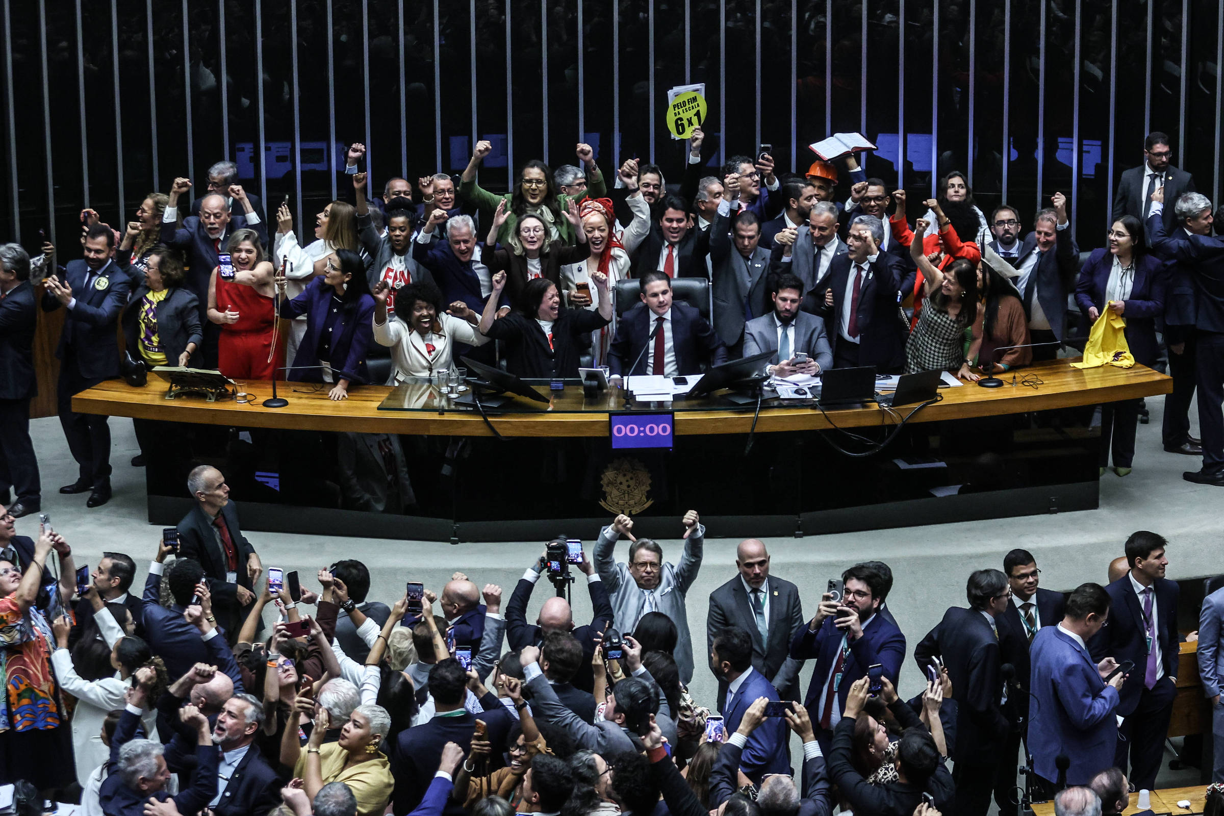 Grupo numeroso de deputados em pé atrás da mesa principal do plenário, muitos com os punhos erguidos em comemoração. Na frente, público e outros parlamentares também registram o momento com celulares. Ambiente interno com iluminação artificial e estrutura típica de assembleia legislativa.
