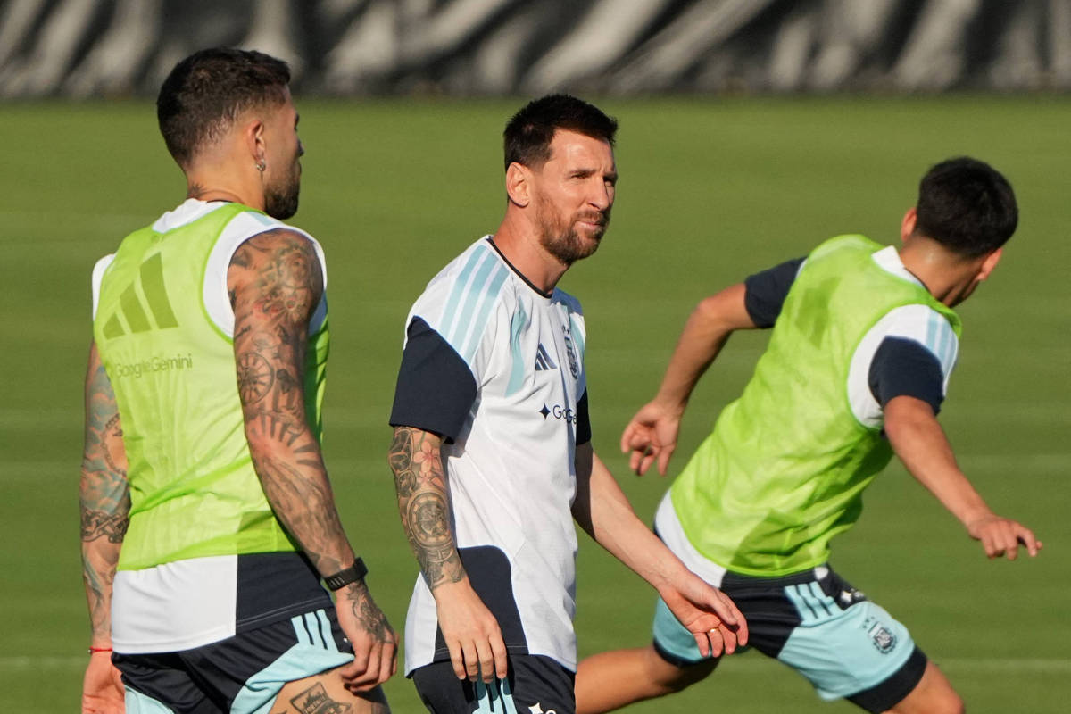 Três jogadores de futebol em campo de grama durante treino. Dois vestem coletes verdes e um camisa branca com detalhes azuis, todos com shorts claros. O jogador central está de perfil, com expressão concentrada, enquanto os outros dois se movimentam ao redor.