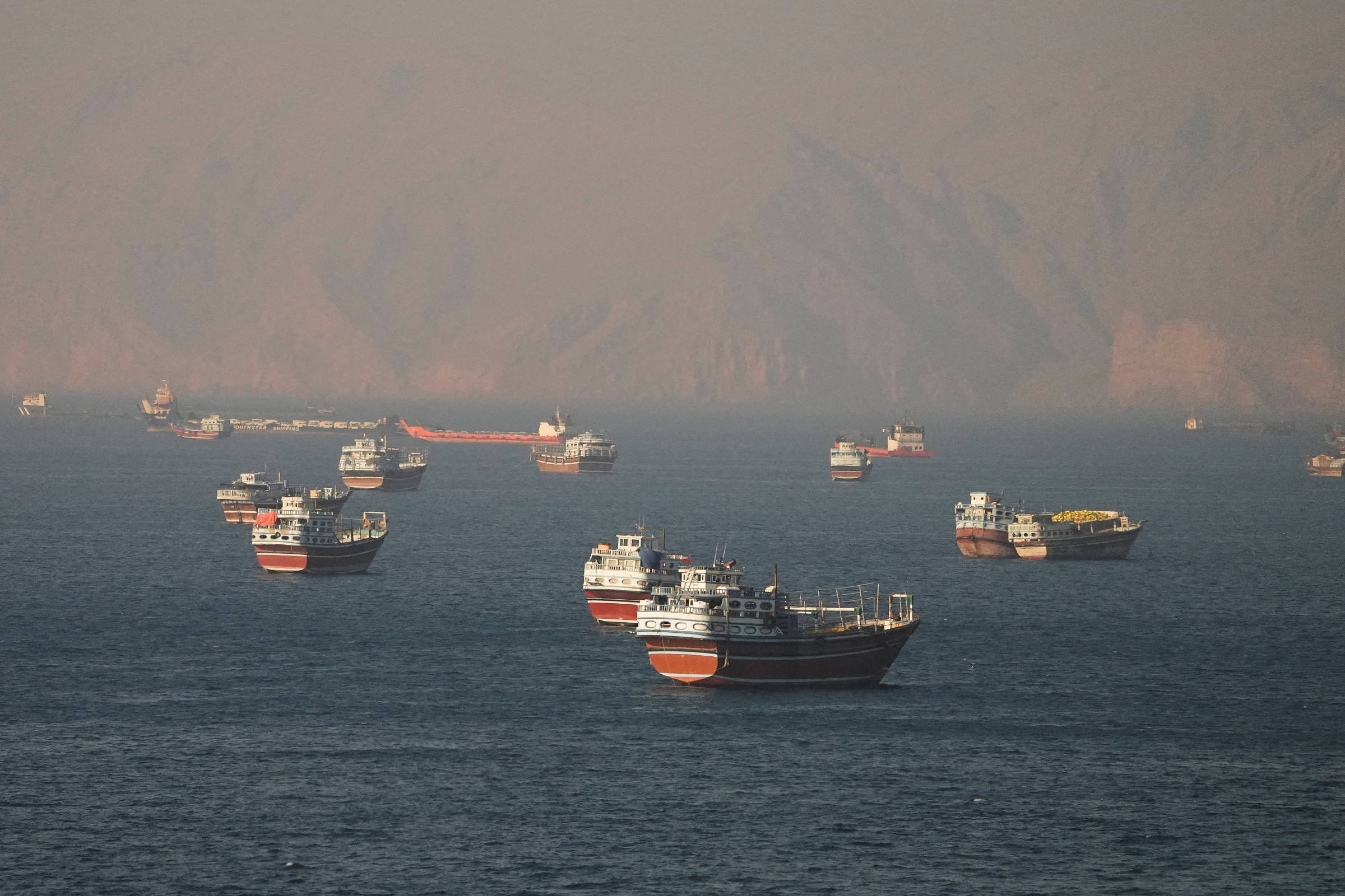 Diversos barcos de pesca ou transporte estão ancorados em águas calmas, distribuídos em diferentes distâncias. Ao fundo, montanhas cobertas por névoa ou fumaça formam um cenário desfocado.