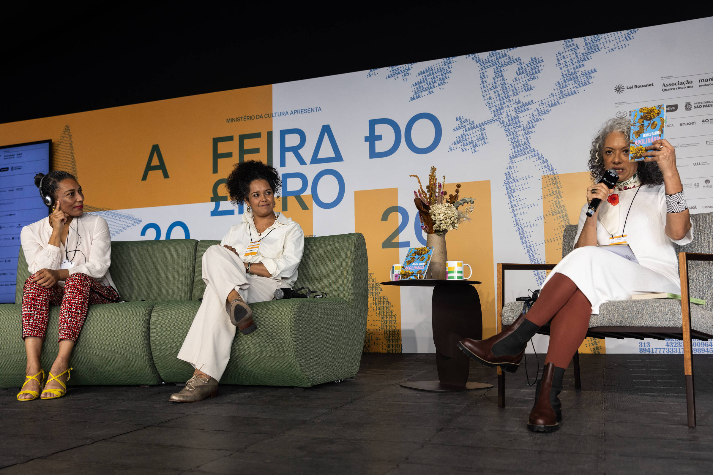 Três mulheres sentadas em palco durante evento da Feira do Livro. Duas estão no sofá verde à esquerda, uma com microfone e outra com fones de ouvido, e a terceira está à direita em cadeira, segurando um livro e falando ao microfone. Ao fundo, painel com texto