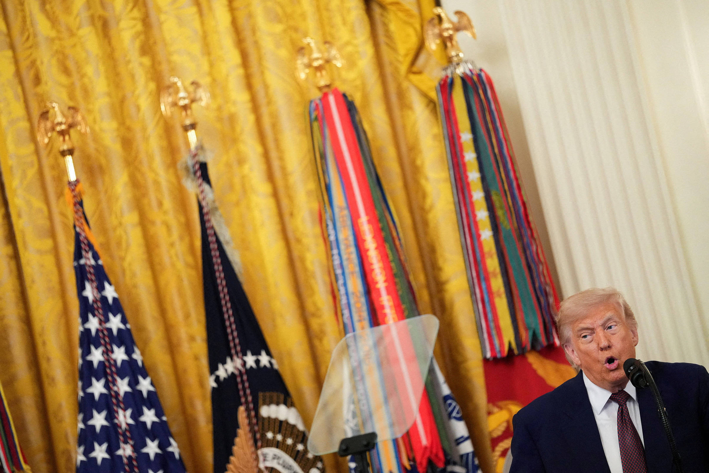Ex-presidente dos Estados Unidos fala em púlpito com microfone, ao lado de quatro bandeiras oficiais, incluindo a bandeira americana e a presidencial, em ambiente interno com cortinas douradas.