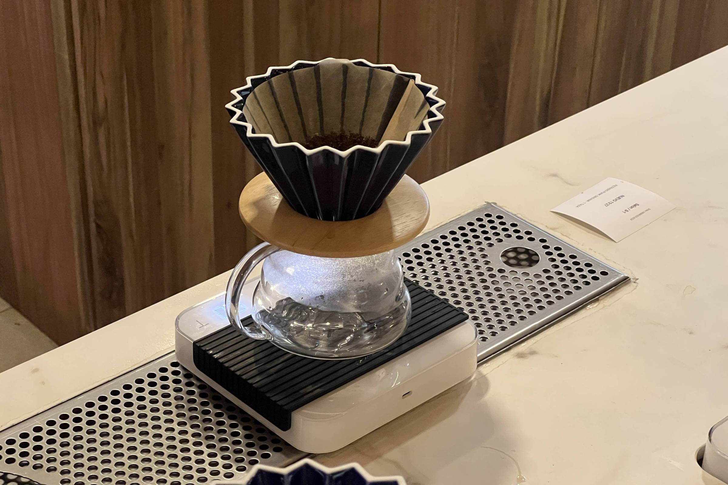 Filtro de papel marrom colocado em suporte preto com base de madeira sobre jarra de vidro transparente. Equipamento está sobre balança digital branca em bancada clara com detalhes metálicos perfurados.