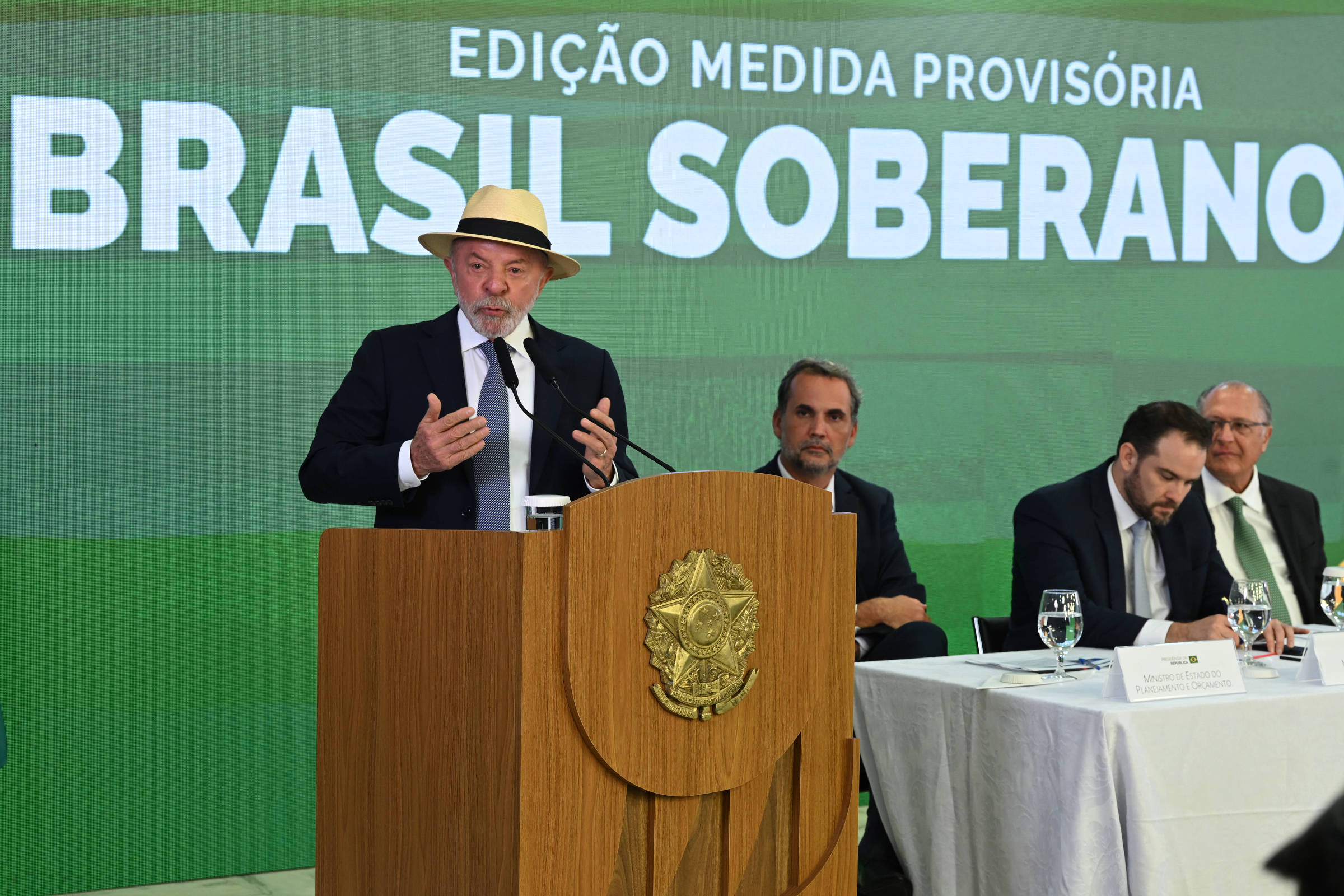 Homem de terno escuro e chapéu claro fala em púlpito com brasão do Brasil. Três homens sentados à direita, um deles escreve. Fundo verde com texto