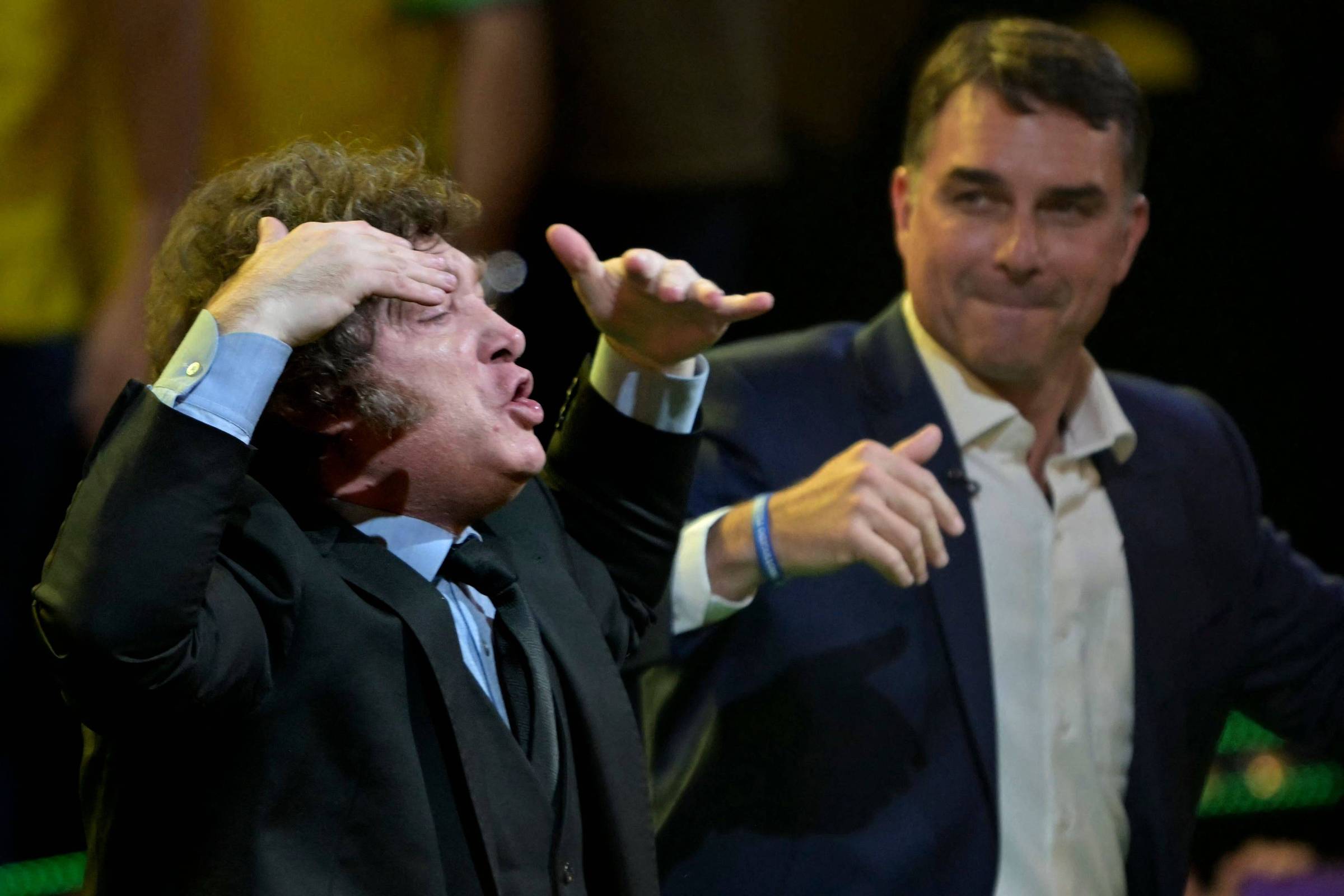 Dois homens em traje formal participam de evento. Homem à esquerda gesticula com as mãos próximas à cabeça, enquanto homem à direita observa com leve sorriso e mão levantada.