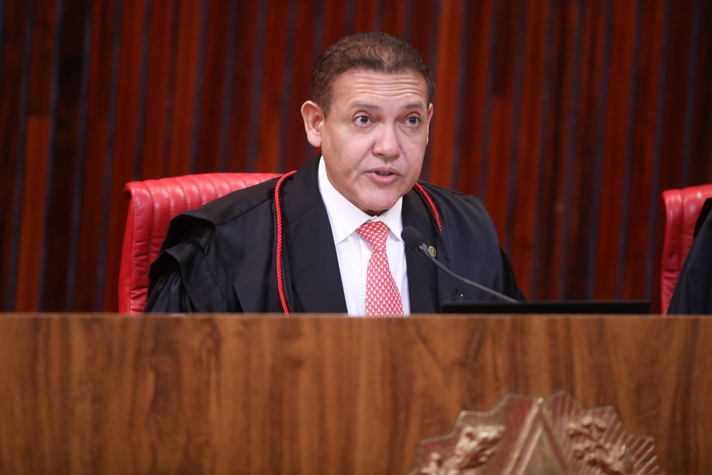 Homem de meia-idade veste toga preta com detalhes vermelhos e gravata vermelha, sentado em cadeira vermelha atrás de bancada de madeira em ambiente formal com painéis de madeira ao fundo.