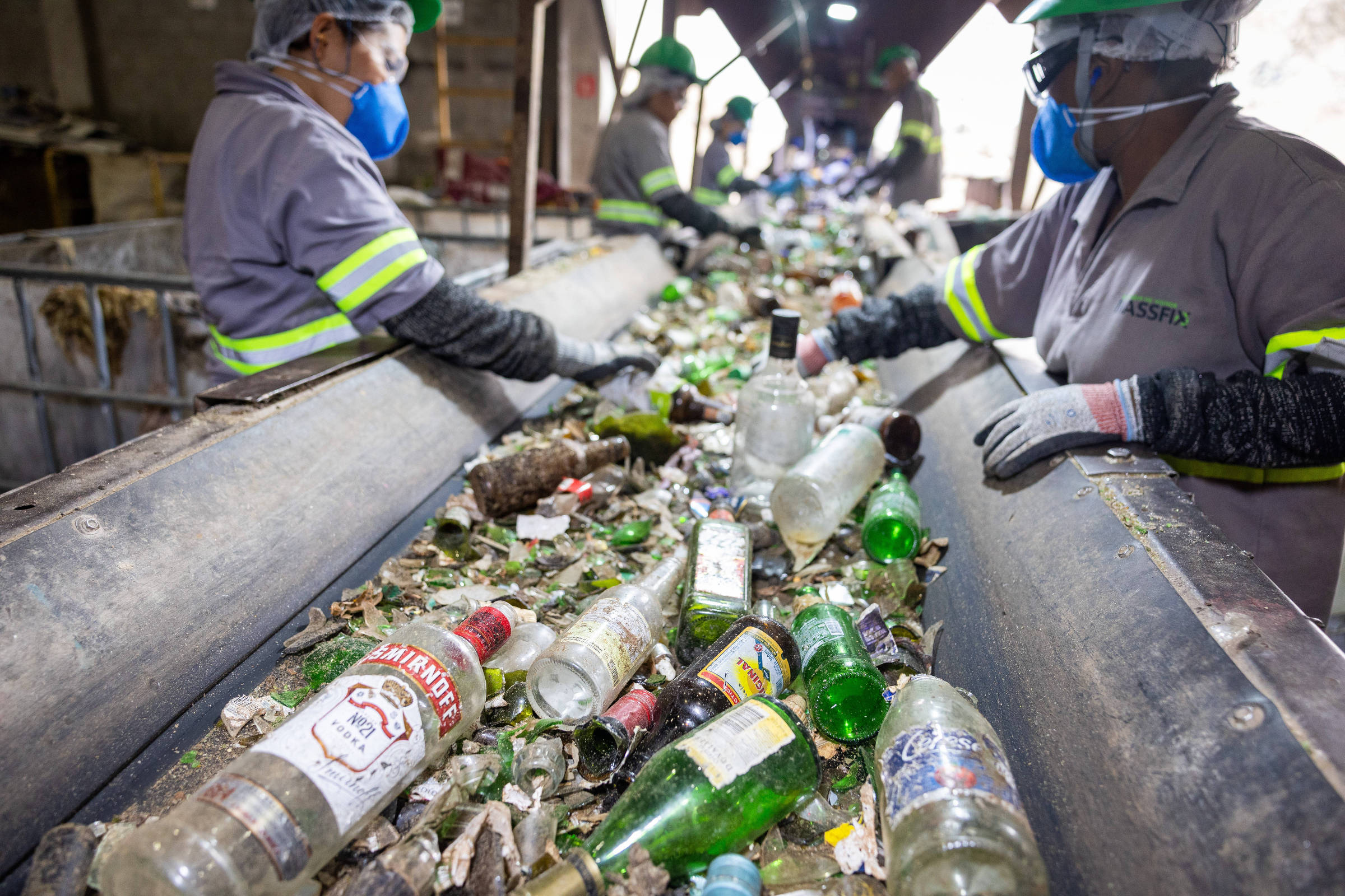 Quatro trabalhadores com uniformes cinza e capacetes verdes separam garrafas de vidro em esteira de triagem em ambiente industrial. Garrafas transparentes e verdes, além de cacos de vidro, estão espalhados na esteira.