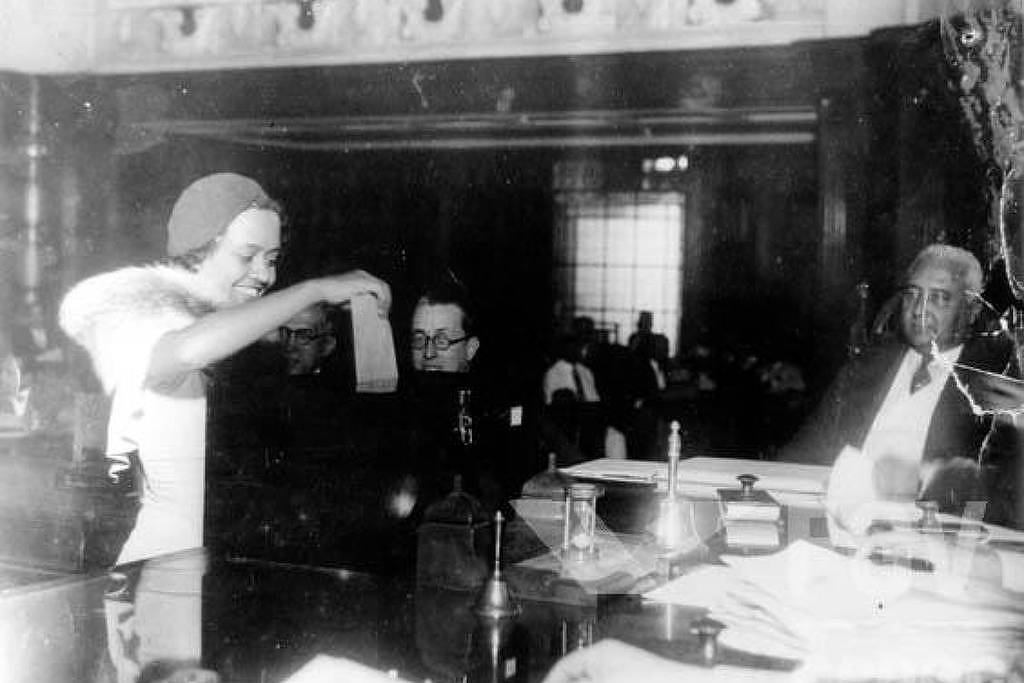 A imagem mostra uma cena de tribunal, onde uma mulher com um vestido claro e um chapéu está em pé, levantando um documento. À sua frente, um homem de óculos está sentado, enquanto outro homem mais velho, com cabelo grisalho, observa a cena. O ambiente é formal, com mesas e documentos visíveis, e há várias pessoas sentadas ao fundo, assistindo ao que acontece.