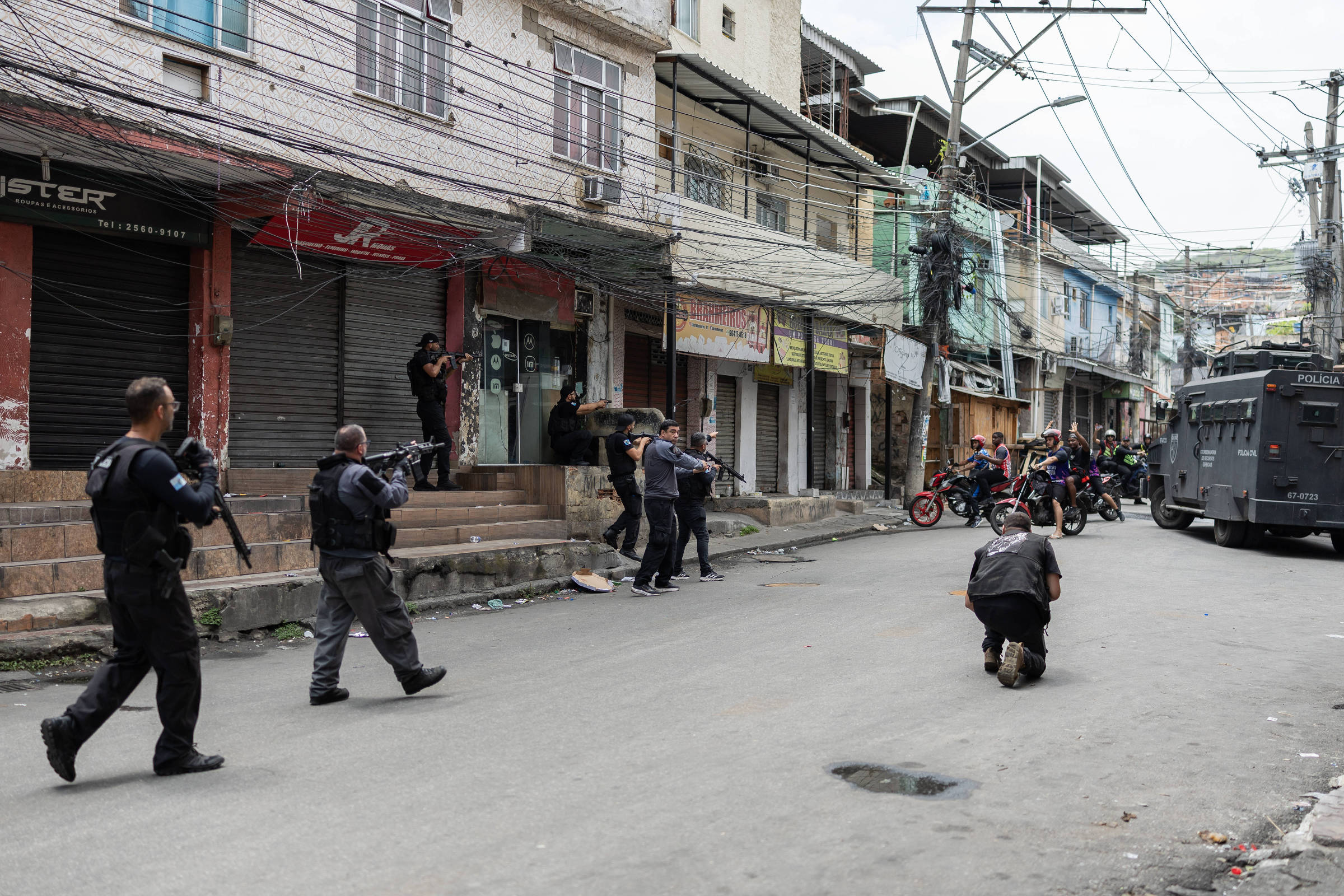 Seis policiais armados avançam por rua com prédios comerciais fechados. Um policial está ajoelhado no meio da rua, enquanto outros se aproximam de uma entrada. Ao fundo, grupo de pessoas observa próximo a motocicletas e viatura blindada estacionada.