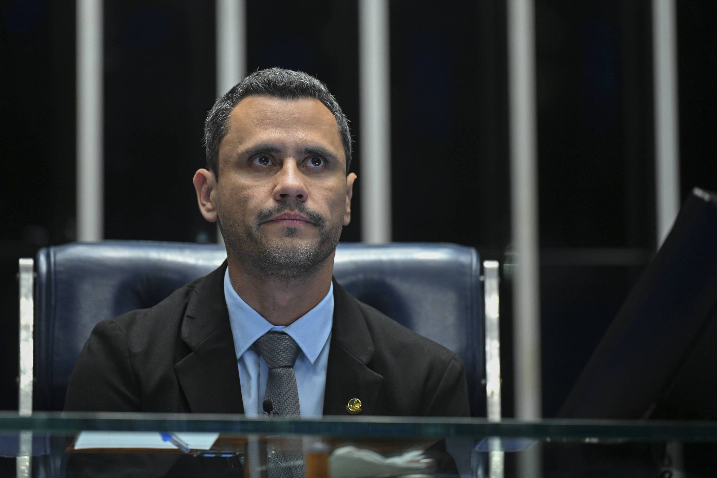 Homem de cabelos curtos e escuros veste terno preto, camisa azul clara e gravata cinza, sentado em cadeira de couro azul escuro, com fundo de painéis verticais pretos e brancos.