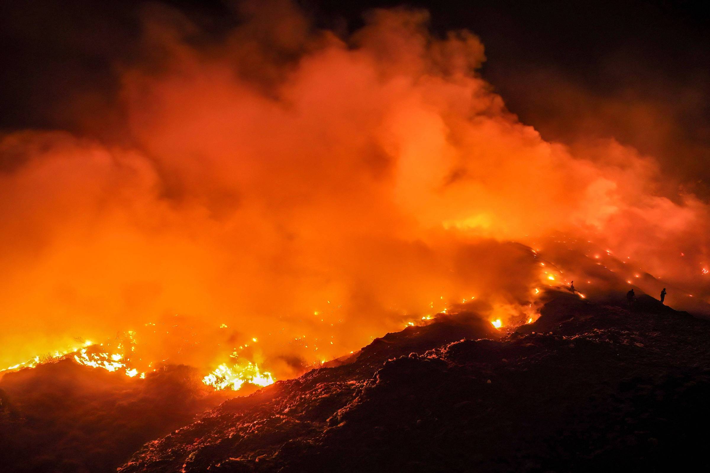 Fogo intenso consome vegetação em área de serra durante a noite, com grandes nuvens de fumaça alaranjada e chamas visíveis ao longo do terreno irregular.