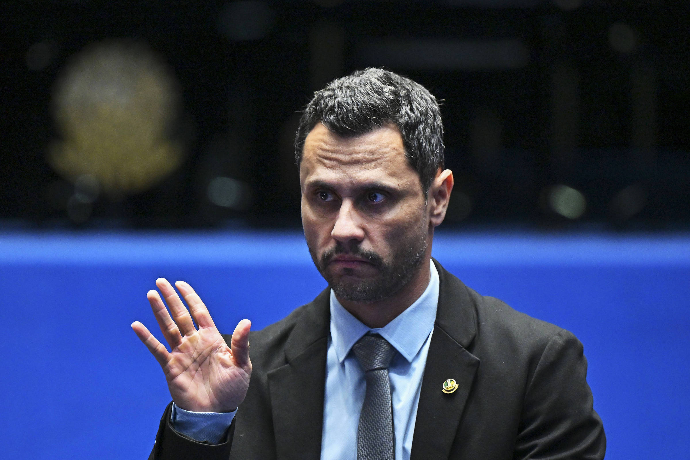 Homem de cabelos curtos e escuros, vestido com terno preto, camisa azul clara e gravata cinza, levanta a mão direita em gesto de saudação ou juramento. Fundo desfocado em azul com símbolo oficial dourado.