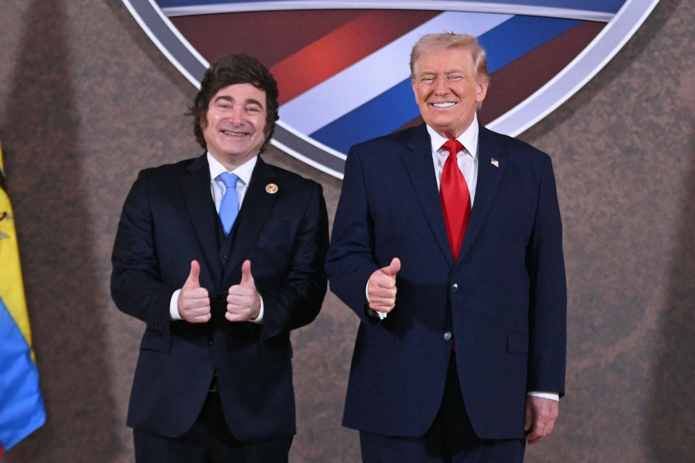 Dois homens de terno escuro, um com gravata azul clara e outro com gravata vermelha, posam lado a lado sorrindo e mostrando polegares para cima. Ao fundo, parte de um escudo com cores azul, branco e vermelho é visível.