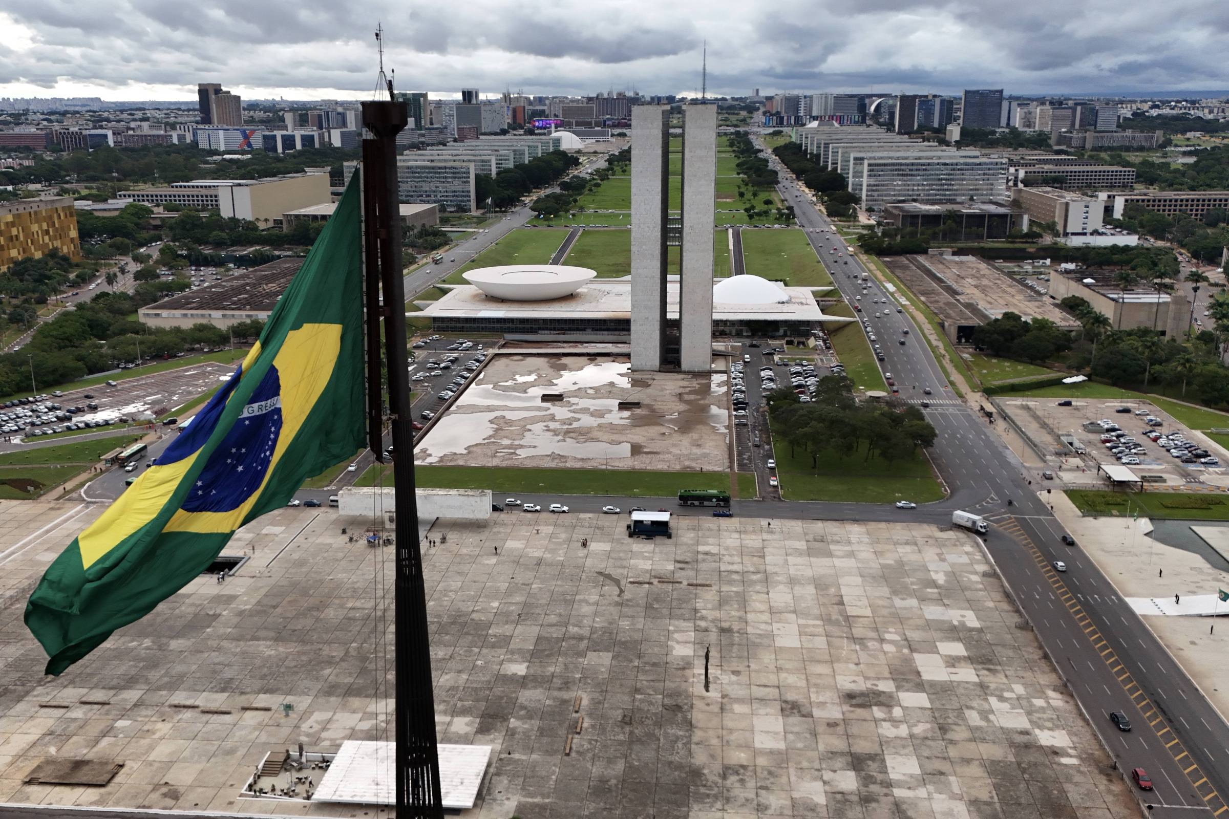 A imagem mostra uma vista panorâmica da Esplanada dos Ministérios em Brasília, com a bandeira do Brasil em primeiro plano. Ao fundo, destacam-se os edifícios do Congresso Nacional e o Palácio da Alvorada, além de uma ampla área verde e ruas. O céu está nublado, sugerindo um clima encoberto.