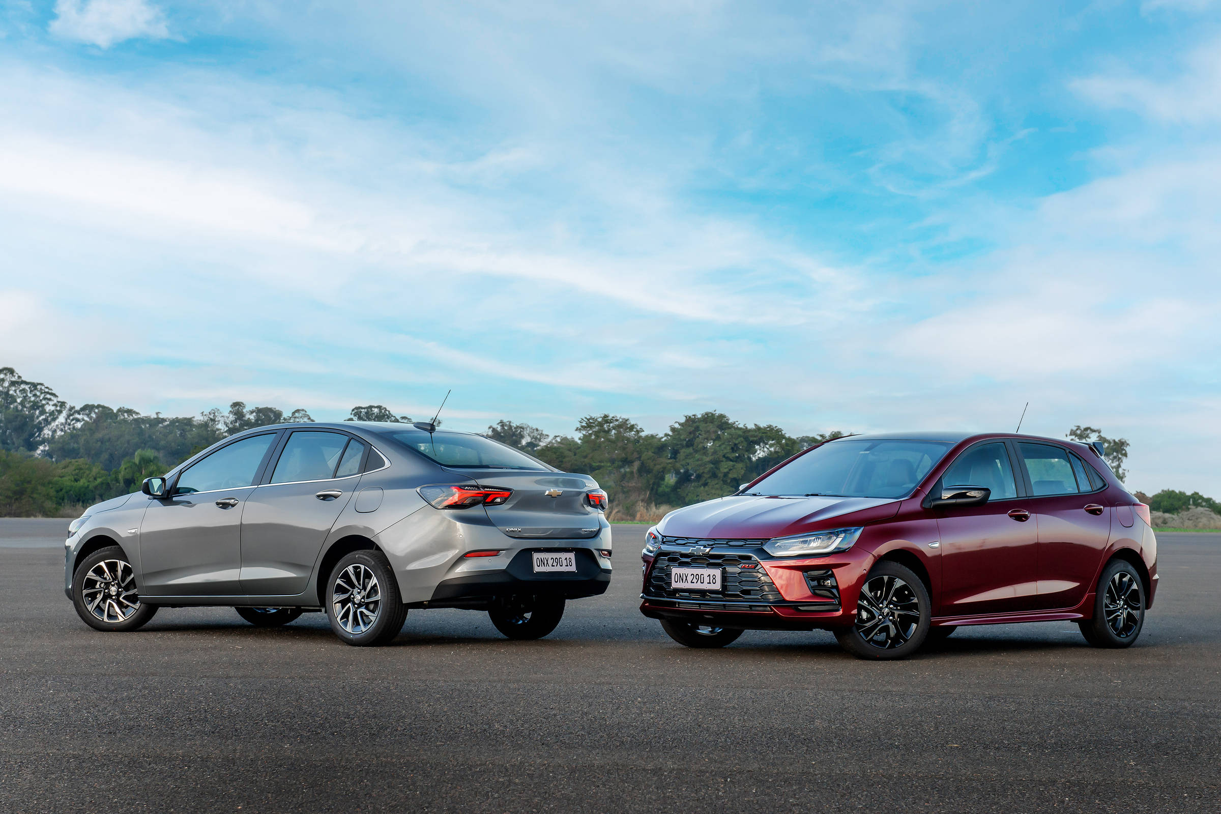 A imagem mostra dois carros estacionados lado a lado em uma pista. O carro à esquerda é de cor prata e tem um design sedã, enquanto o carro à direita é de cor vermelha e possui um design hatchback. O céu está claro com algumas nuvens e há vegetação ao fundo.