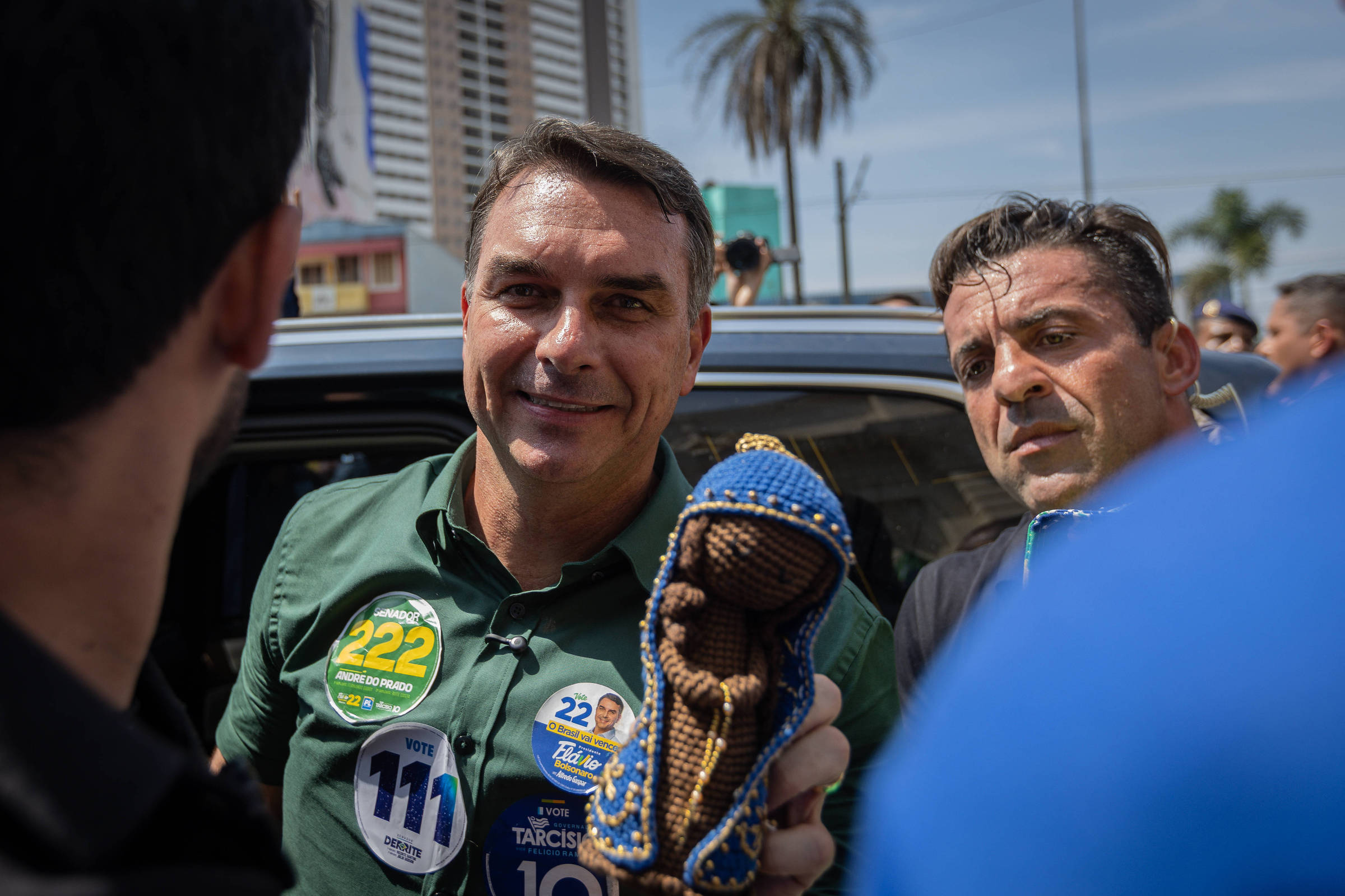 Homem de camisa verde segura uma imagem religiosa azul e dourada enquanto conversa com outras pessoas em ambiente externo, com prédios e palmeira ao fundo.