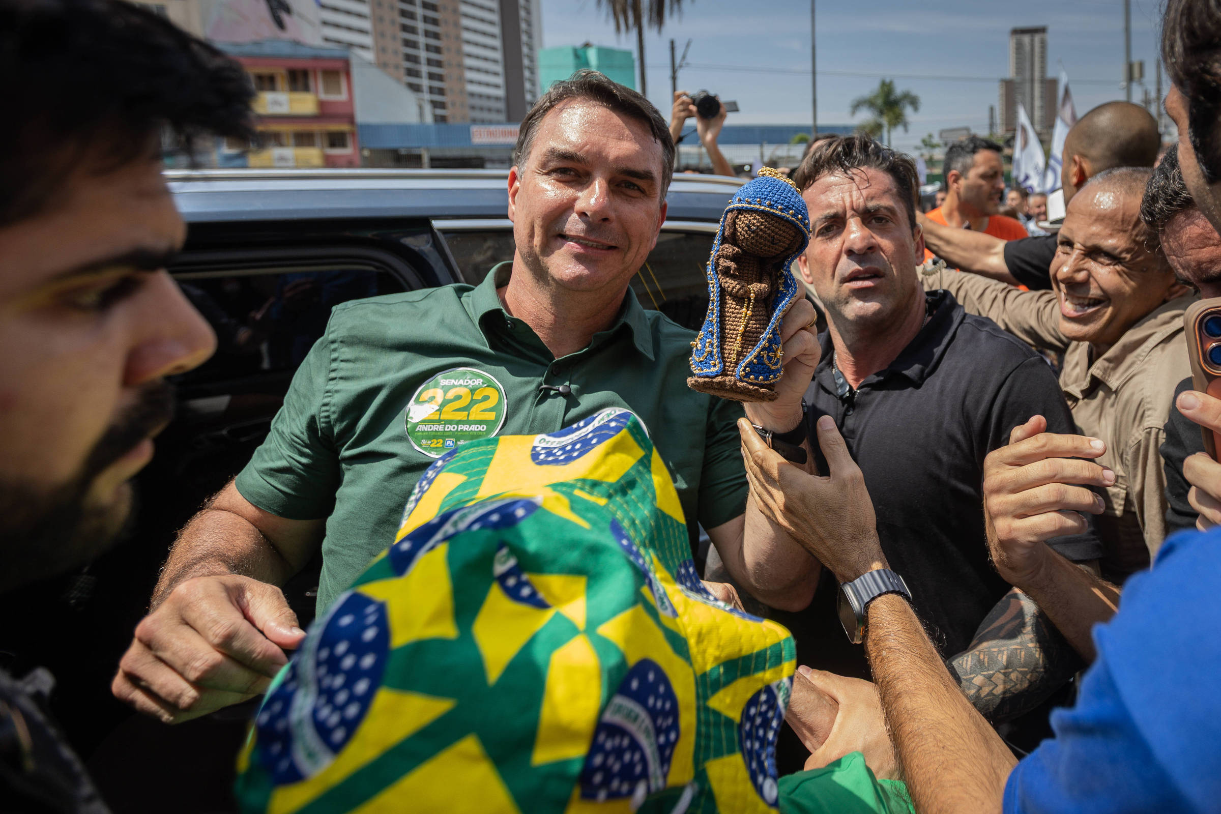 Homem com camisa verde e adesivo de campanha é cercado por grupo de pessoas em área urbana. Um dos presentes segura uma escultura pequena de figura humana.