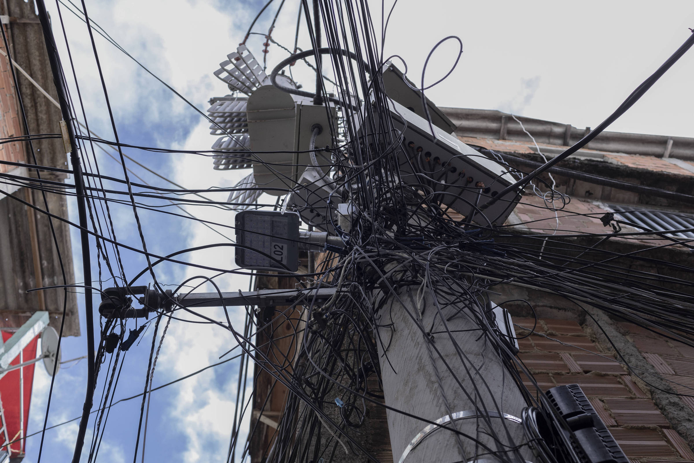Poste de concreto com grande quantidade de fios elétricos e de telecomunicações entrelaçados e desorganizados, conectados a equipamentos e caixas instaladas no poste. Edifícios residenciais aparecem ao fundo sob céu parcialmente nublado.